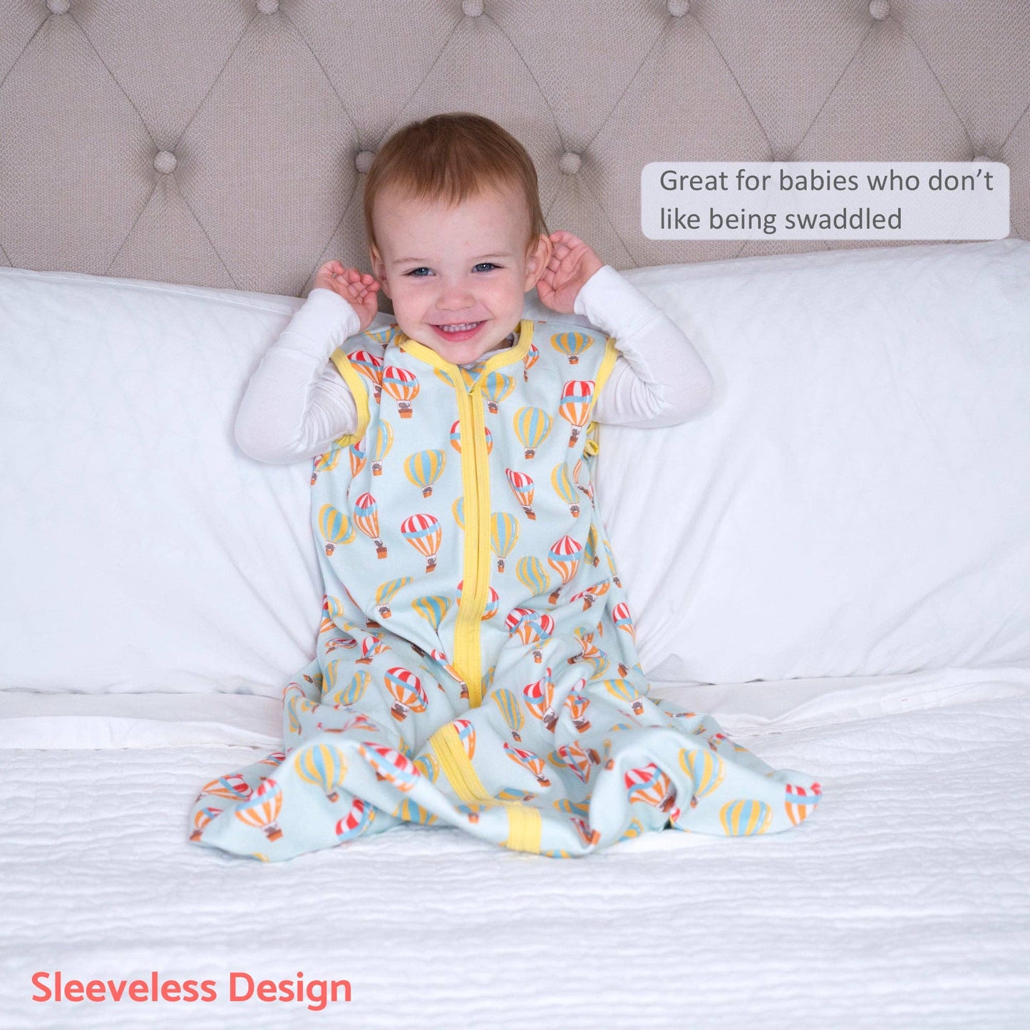 Child wearing a sleeveless sleeping sack with hot air balloon pattern on a bed.