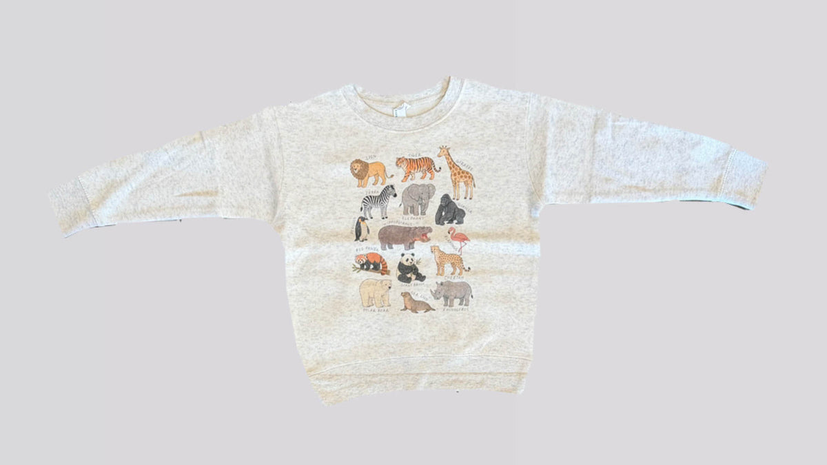A child's heather oatmeal sweatshirt is laid out on a beige background. The sweatshirt has drawn safari animals identified with handwritten text next to each animal. 