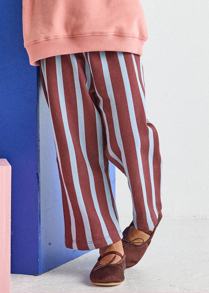 A person wearing loose vertical-striped pants in rust red and light blue, paired with a dusty pink sweatshirt and brown flat shoes, standing next to a blue block.