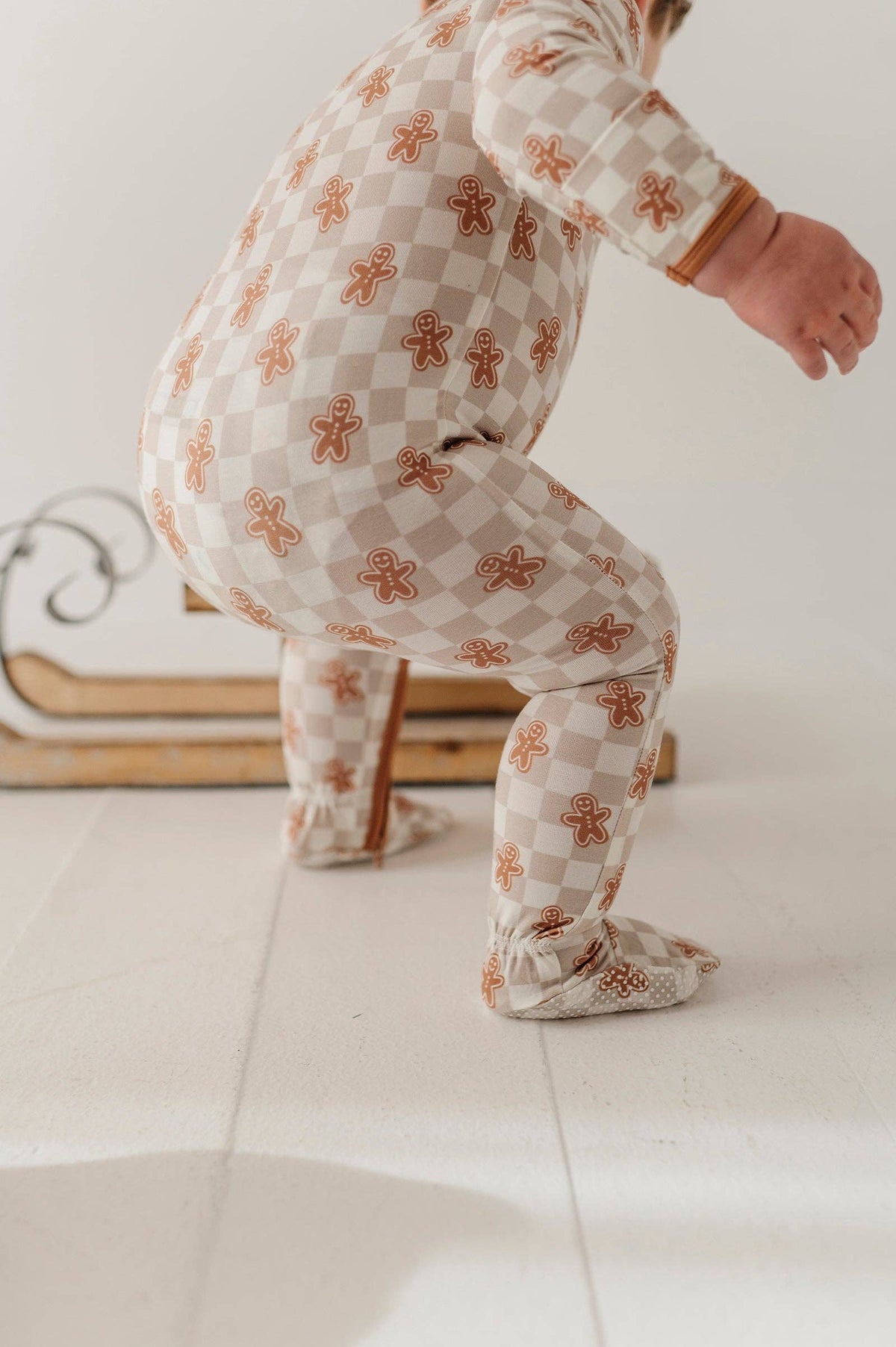 A child wearing a one-piece footed pajama with a beige and white checkerboard pattern and orange-brown gingerbread cookie motifs, crouching on a light floor.