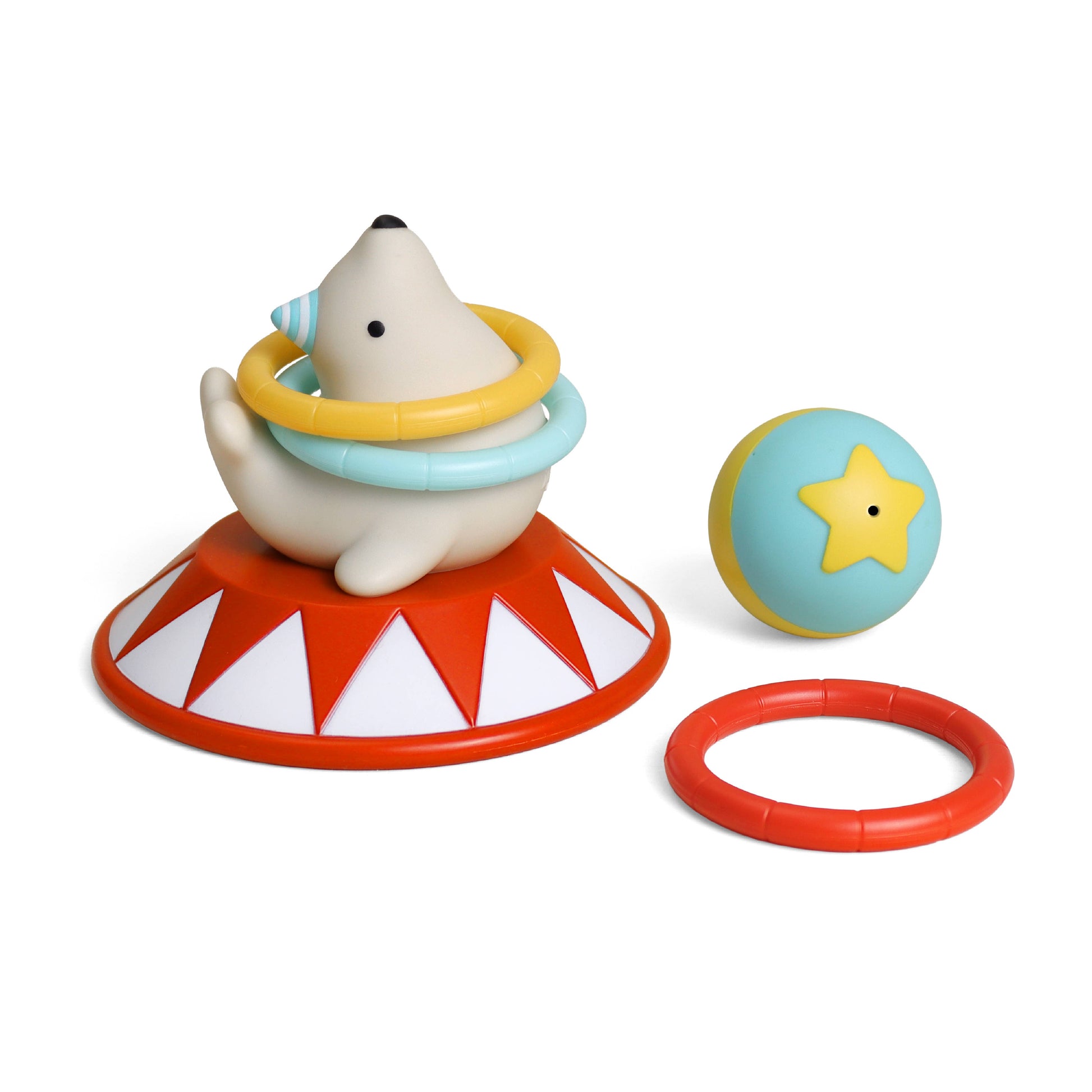 A children's toy set featuring a white seal sitting on a red and white circus-style base, with a yellow and light blue hula hoop around its neck. A light blue ball with a yellow star and a separate red ring are placed beside it on a clean white background.