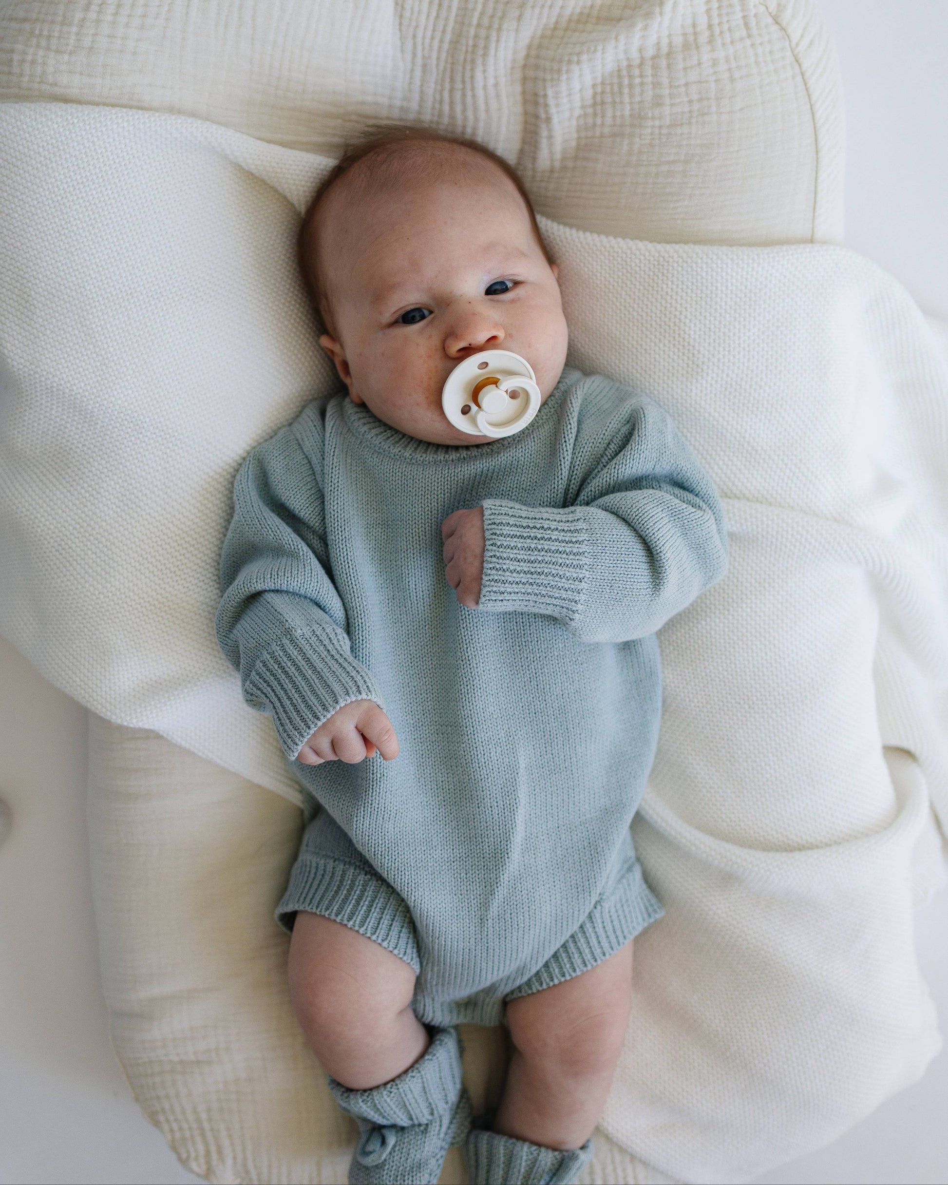 Baby in a light blue knitted outfit lying on a white blanket