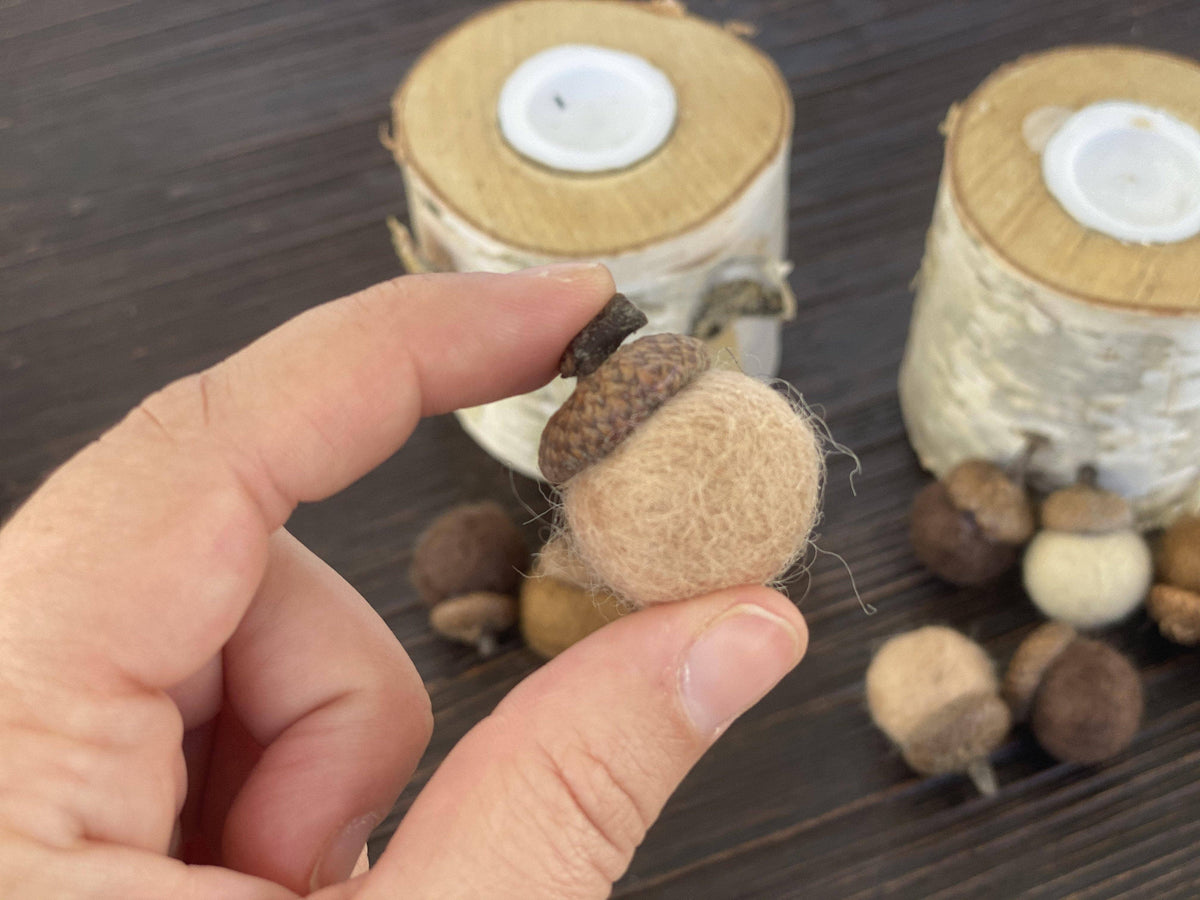 A hand holds a small felted acorn ornament with a brown felted cap and stem, set against a dark wood background with birch log candle holders and additional acorns.