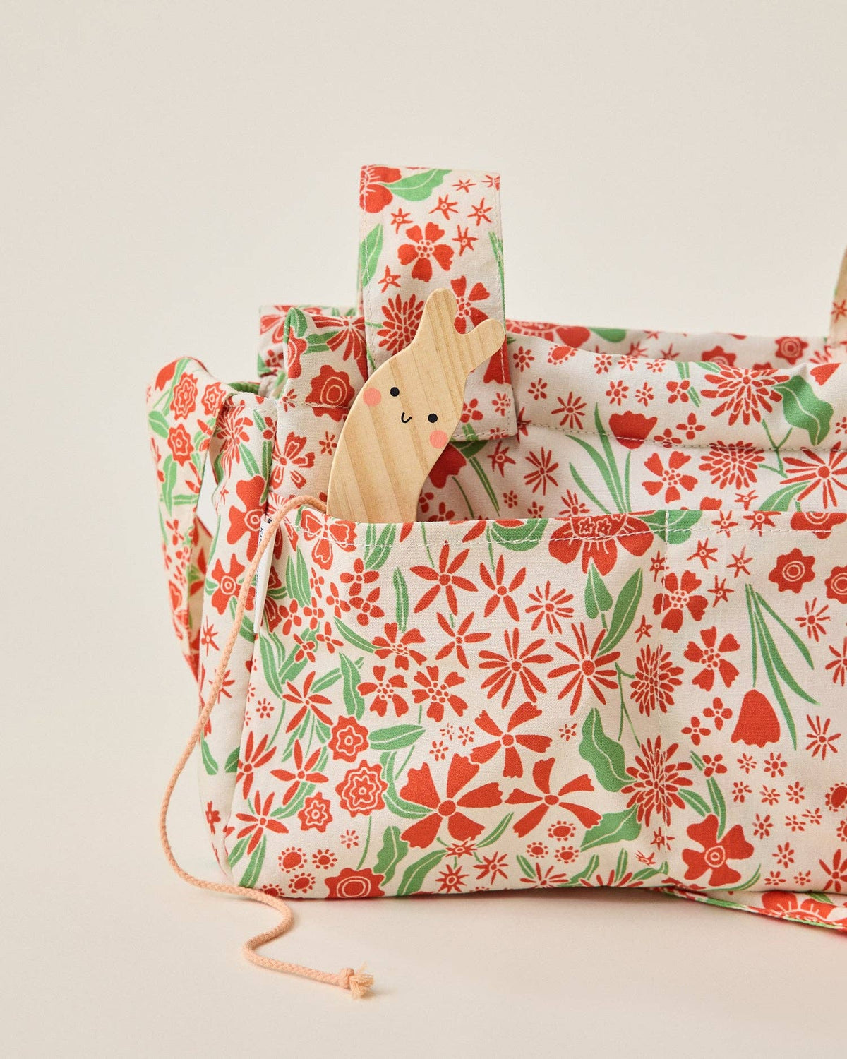 A fabric stroller bag with a red and green floral pattern on a white background, featuring a wooden smiling snail toy tucked into one of its pockets.
