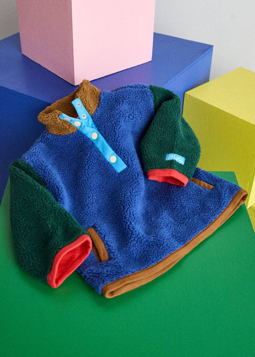 A folded fleece pullover jacket with a blue body, dark green sleeves, brown collar and trim, and red pocket openings, styled on a green surface with colorful geometric blocks in the background.