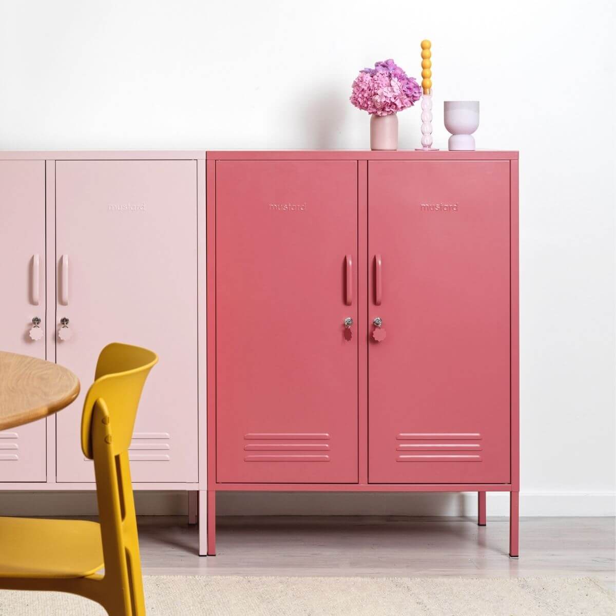 A pink metal two-door storage cabinet with small embossed branding on the doors, standing in a bright room next to a pale pink cabinet and a yellow chair. Decorative pink flowers and small objects sit on top.