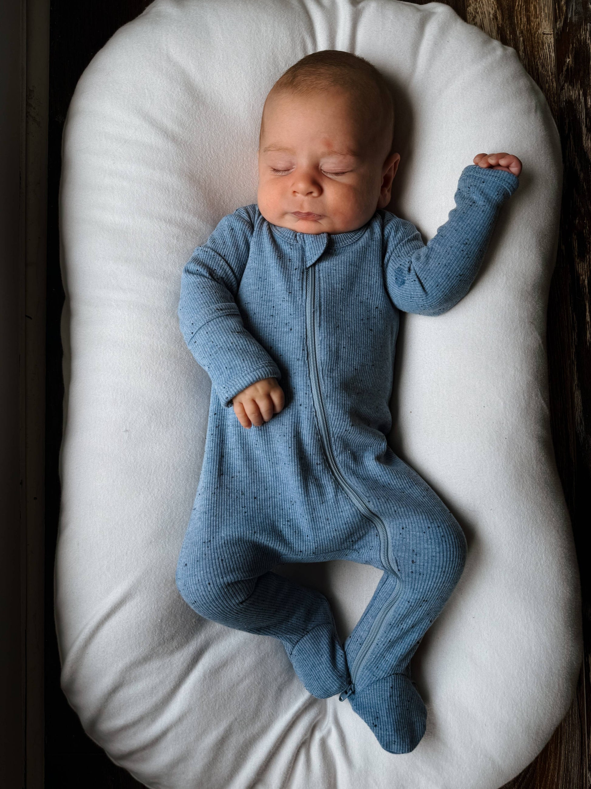 Newborn baby in a blue onesie lying on a white cushion.