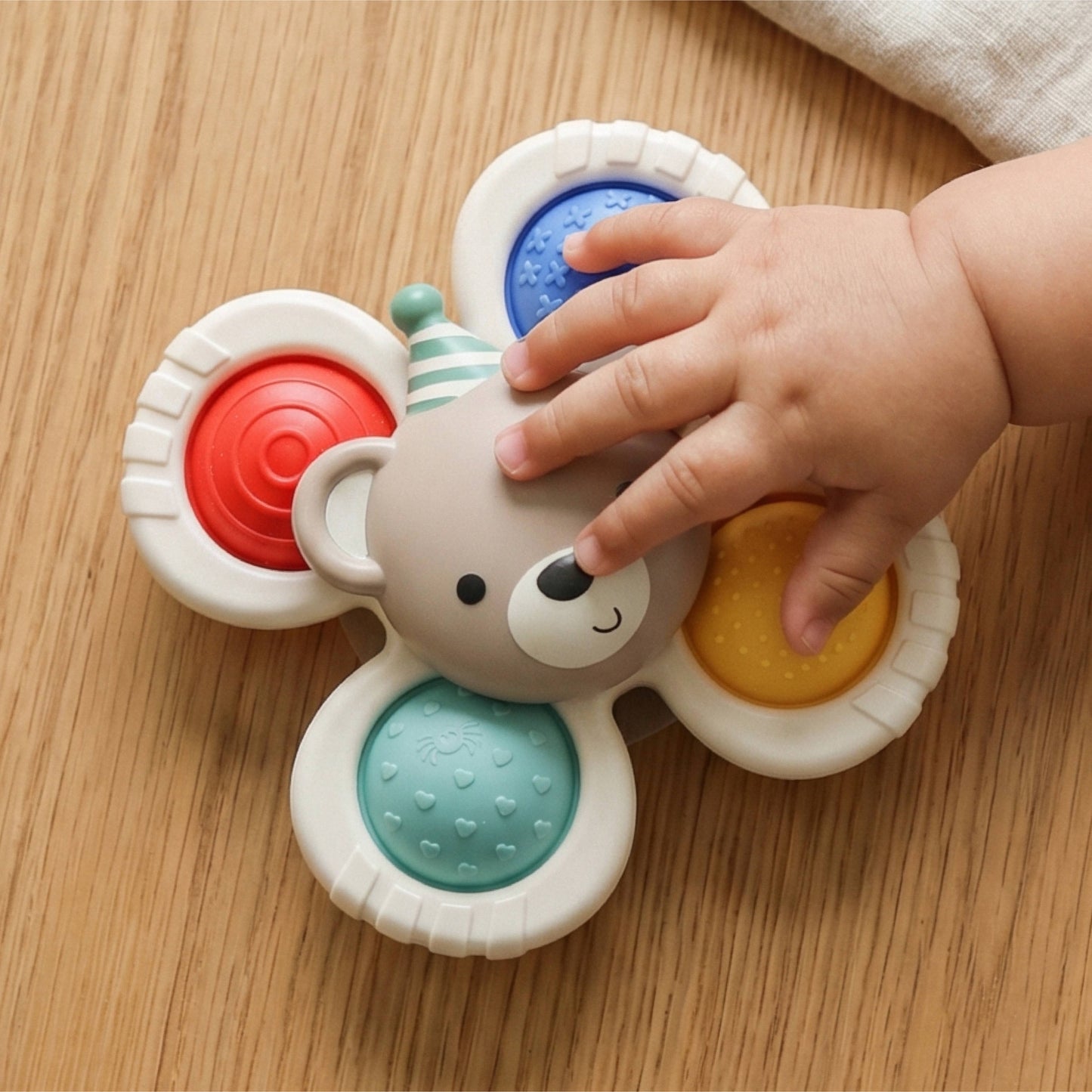 A baby's hand is touching a bear-shaped sensory toy with four colorful textured wheels on a light wood surface.