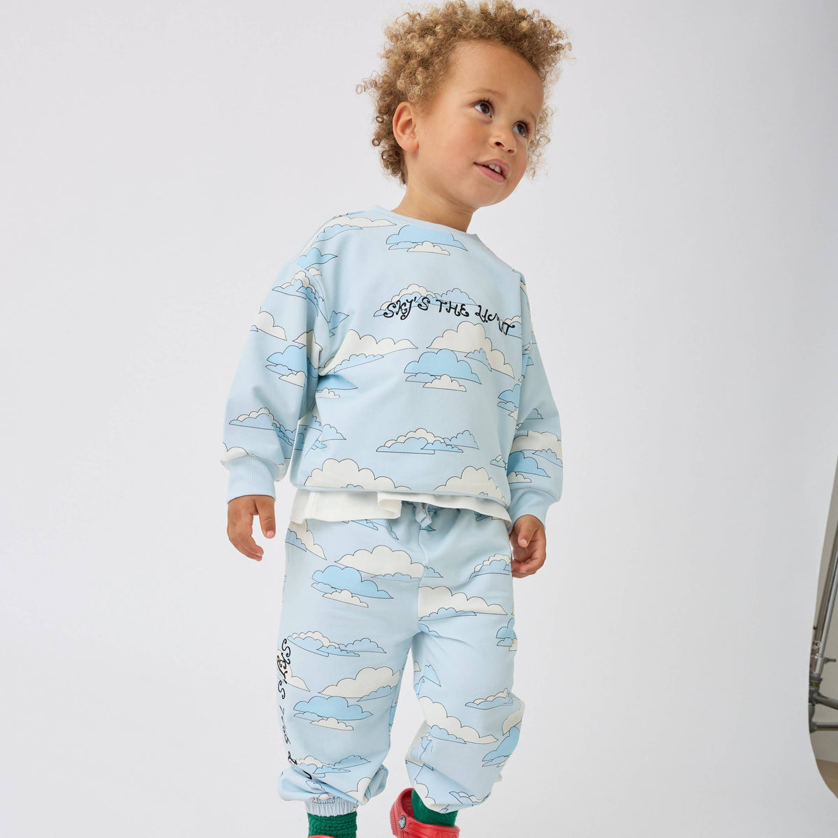 A toddler with curly light brown hair wearing a light blue two-piece pajama set with a white and blue cloud pattern. The long-sleeve top features the text 'Sky's the Limit' in black script, and the matching sweatpants with text on the leg.