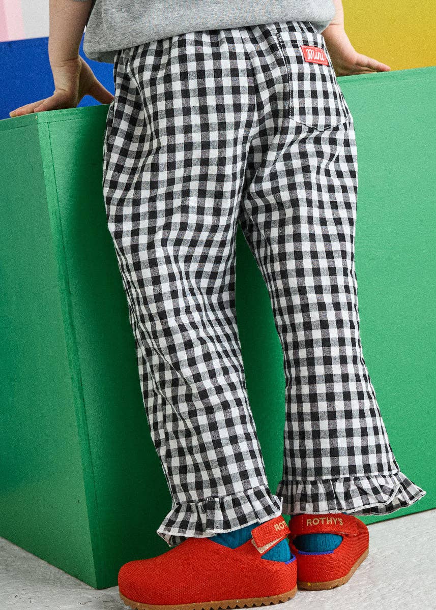 A child wearing black and white gingham check pants with a red brand tab on the back pocket, standing next to a green block.