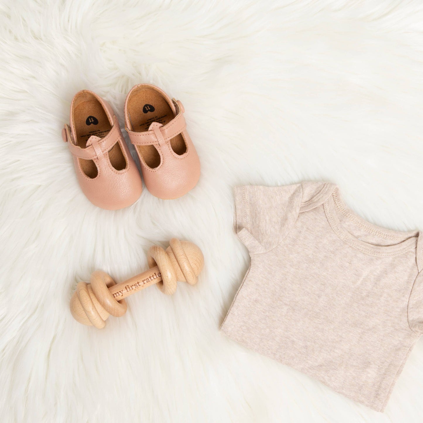 Pink baby shoes, wooden toy, and beige onesie on a white surface