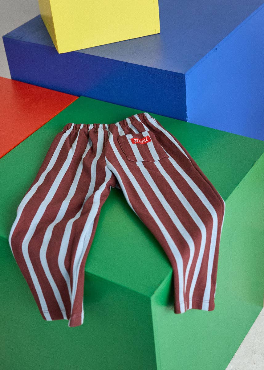 A pair of children's pants with vertical red and white stripes, styled on a green geometric block with colorful background blocks.