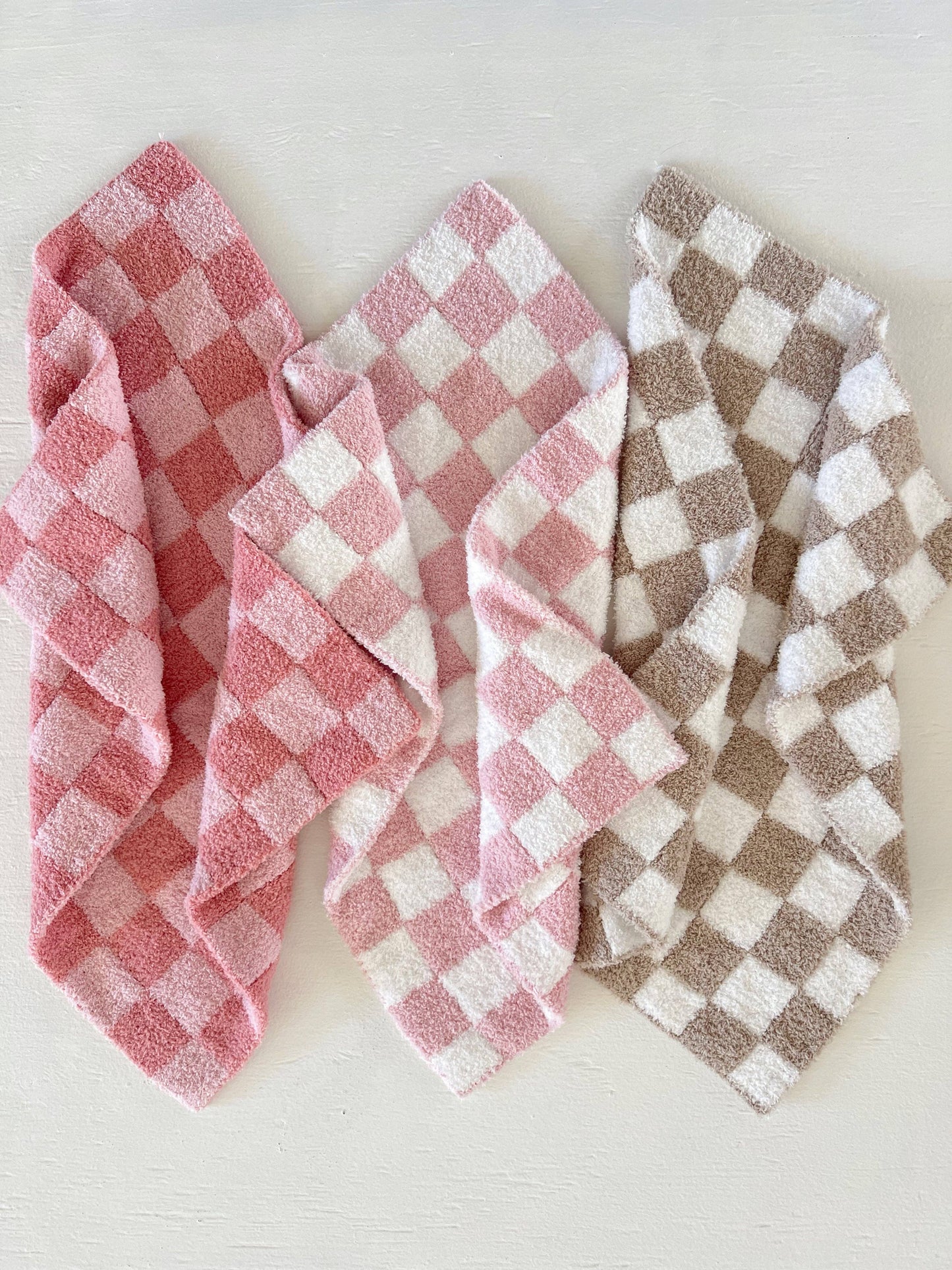 Three checkered baby blankets, one dark and light pink, one pink and white, the other brown and white, on a light background.
