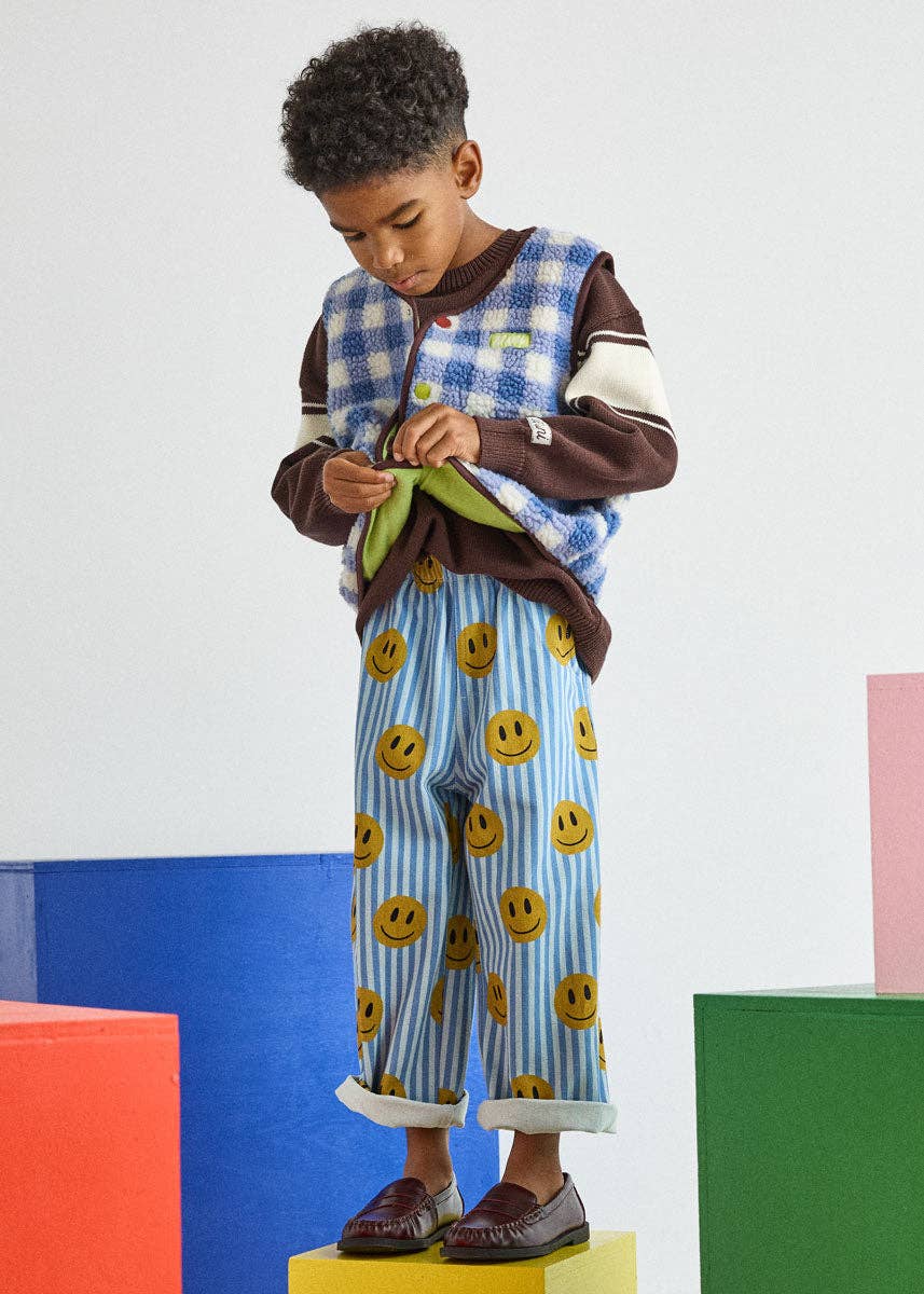 A young boy stands on a yellow block wearing a two-piece pajama set. The set features a brown and blue gingham zip-up vest and light blue and white striped pants with yellow smiley faces.