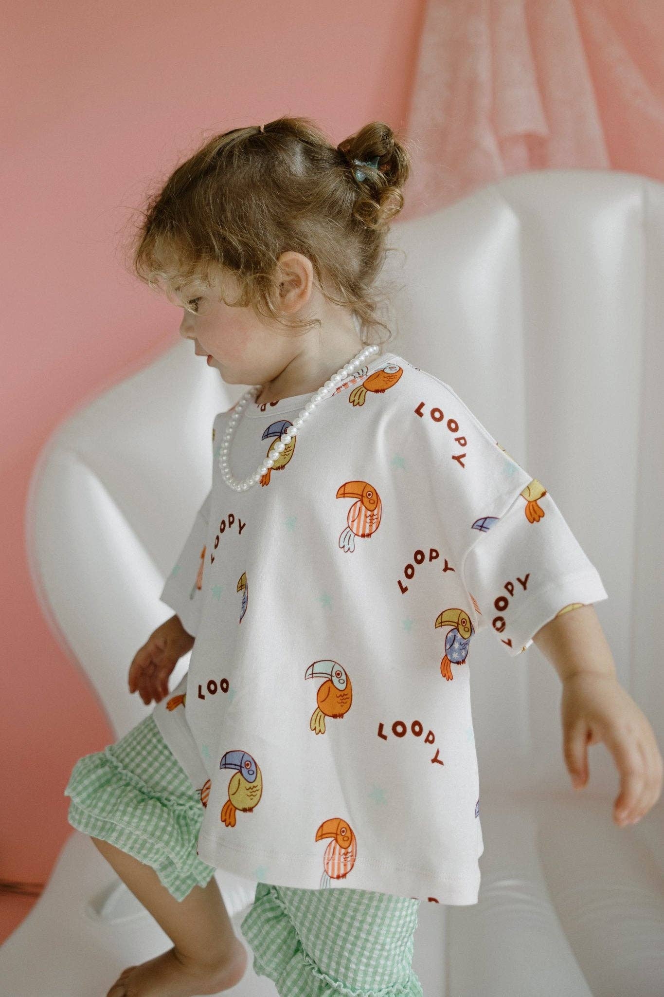 Child wearing a white shirt with colorful patterns and green checkered shorts against a pink background