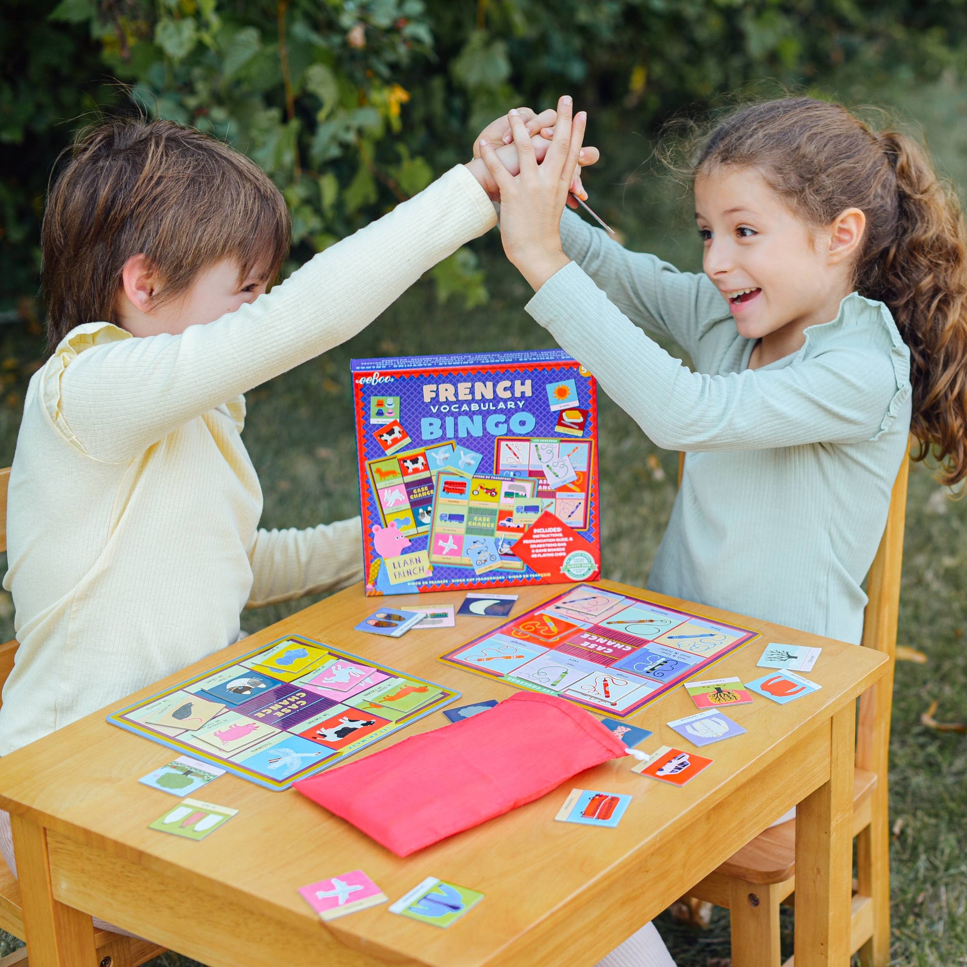 Two children playing with a French vocabulary game at a wooden table outdoors.