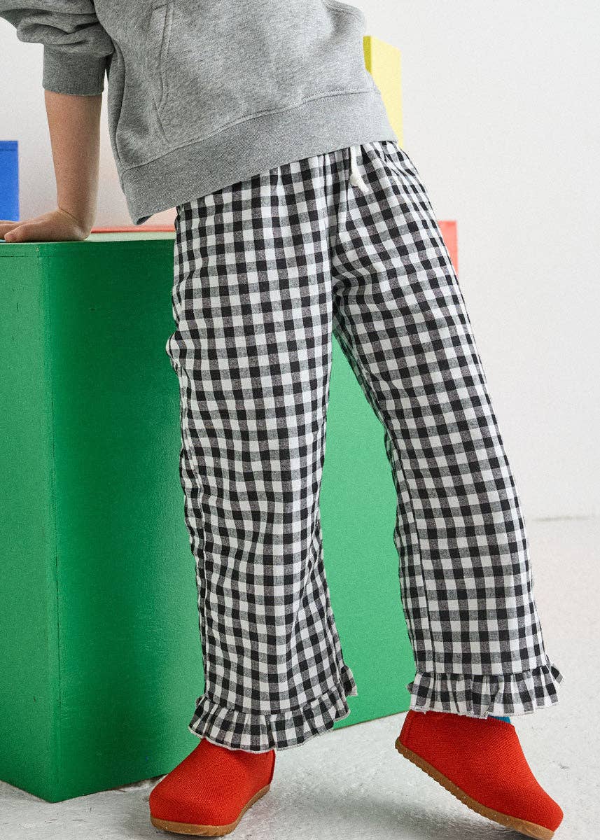 A child wearing black and white gingham wide-leg pants with ruffled hems, paired with a gray hoodie and bright red slip-on shoes, standing next to a green block.