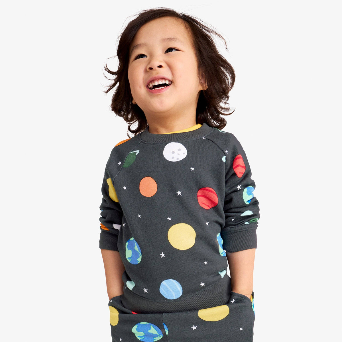 A young child wearing a dark gray long-sleeve sweatshirt and matching pants with a colorful space-themed print of planets, stars, and moons, set against a plain light gray background.
