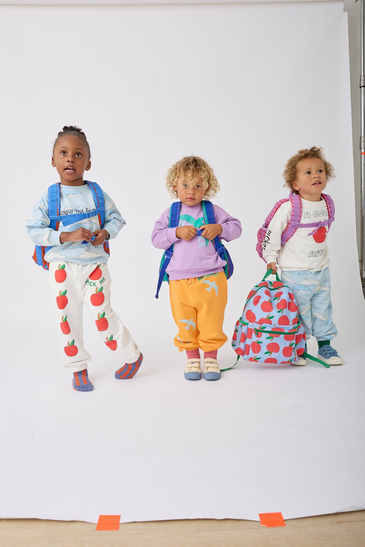 Three young children standing against a white studio backdrop, each wearing a colorful backpack and casual outfit with a red strawberry print. The children are posed together, showcasing the backpacks and matching apparel.