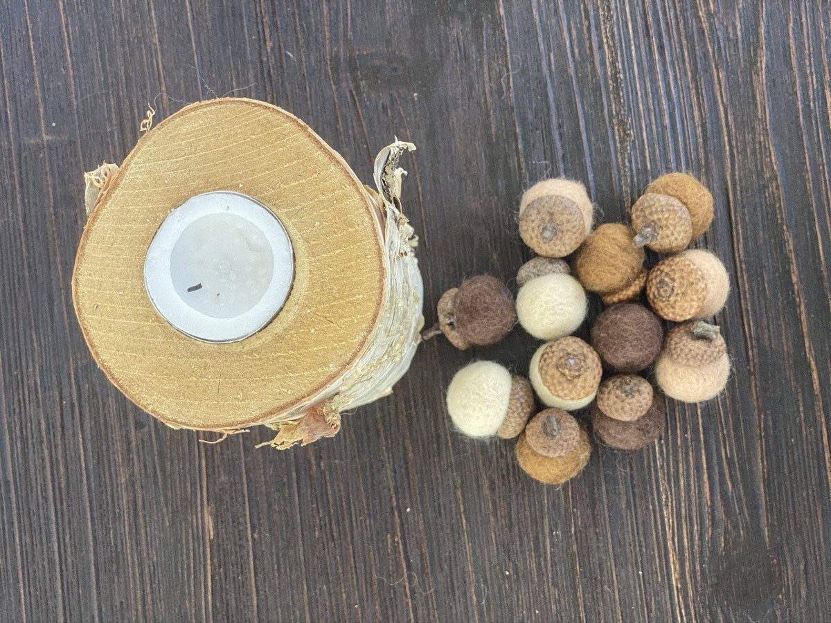 A rustic wooden slice candle with a white wax center sits on a dark wood surface next to a small cluster of dried acorns.