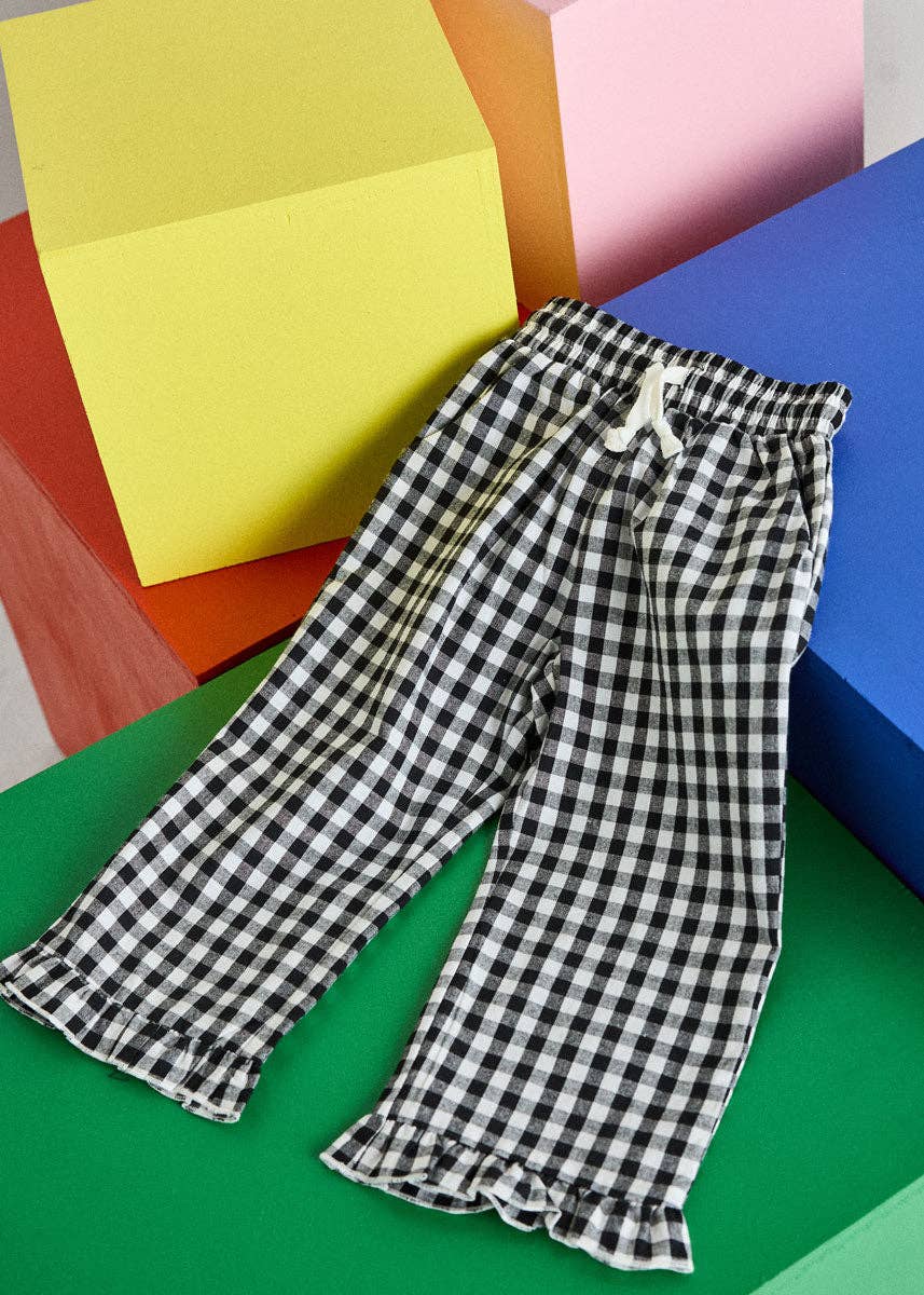 Black and white gingham check cropped pants with a white drawstring waist and ruffled hems, styled on a green surface with colorful geometric blocks in the background.