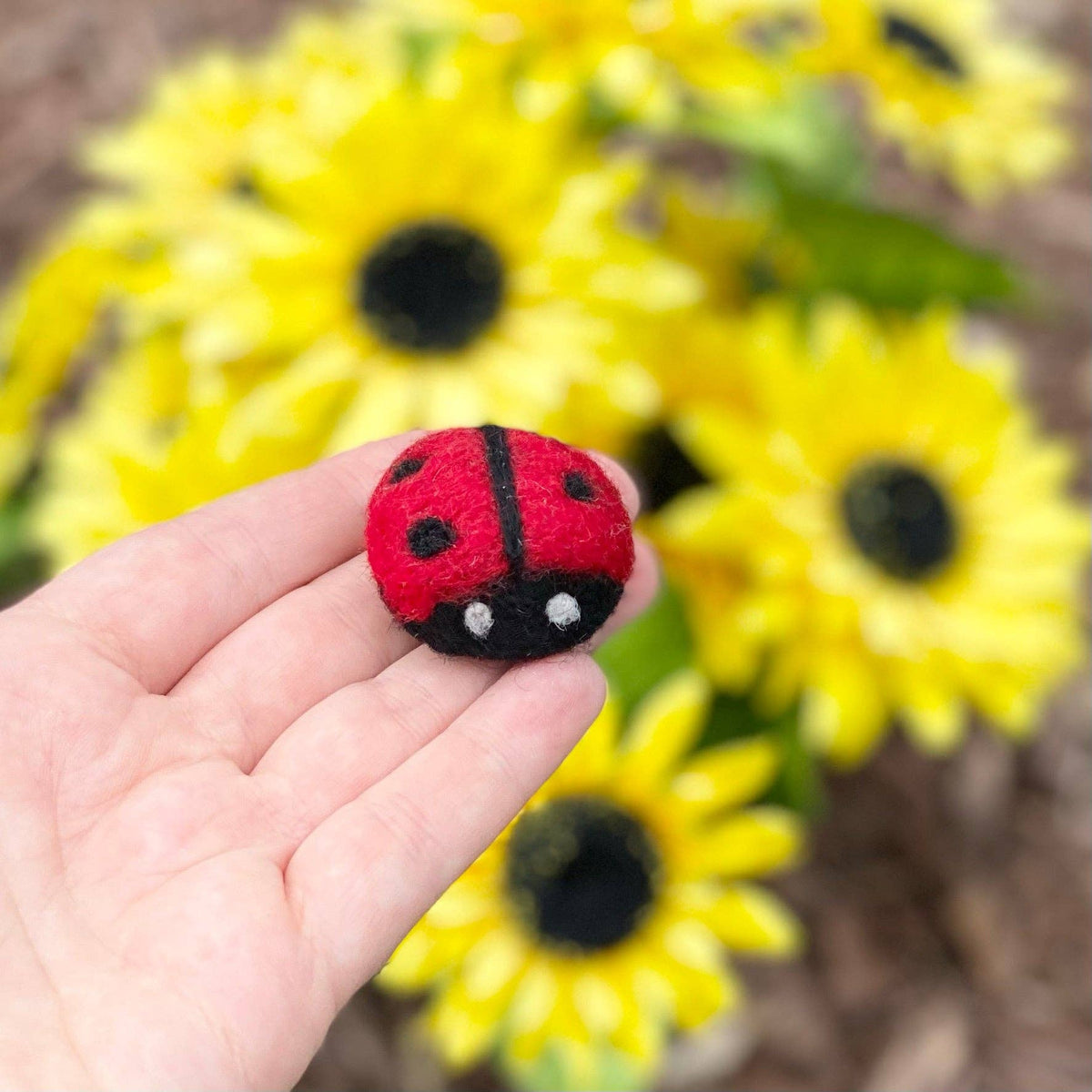 Hand holding a red felt ladybug toy with sunflowers in the background