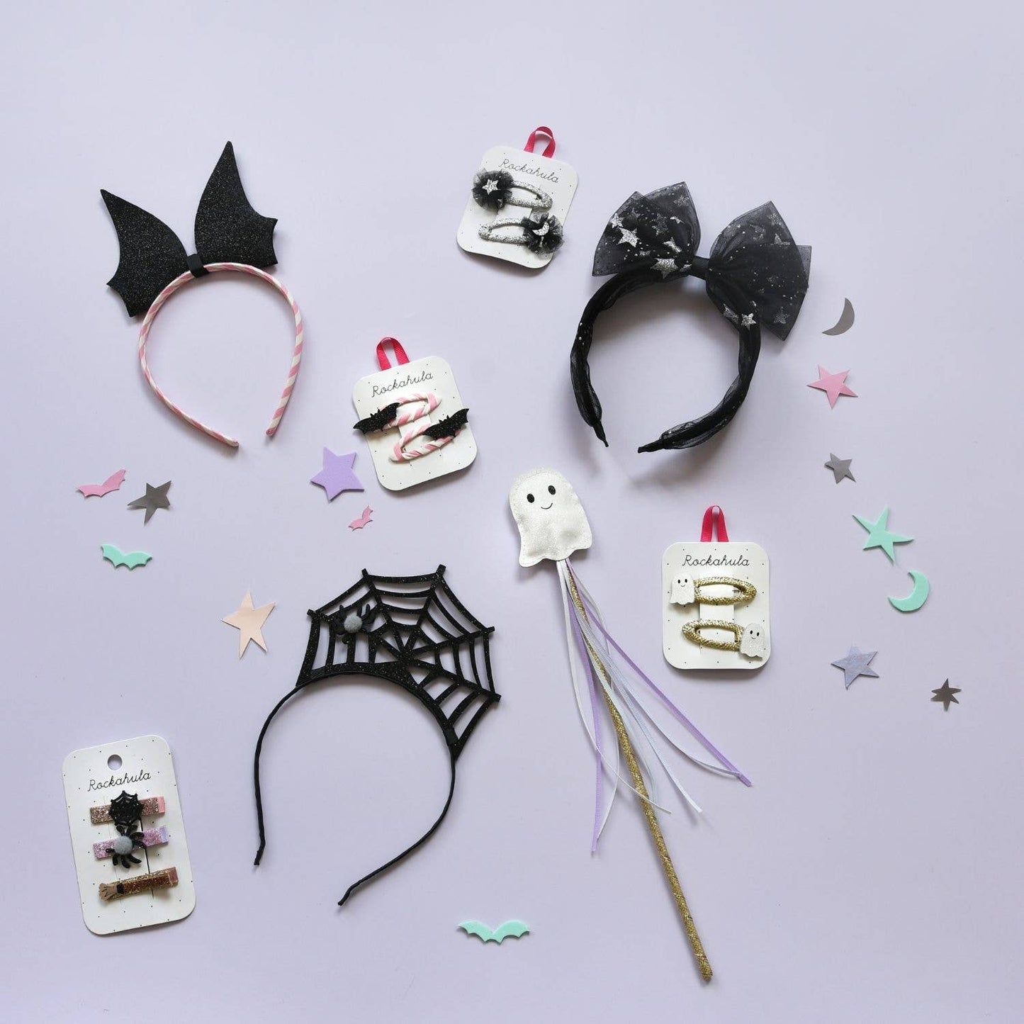 A flat-lay display of various gothic-themed hair accessories on a light lavender background, including black bat ears, a black spiderweb headband, a black bow headband with white glitter, a white ghost hair clip, and other small accessories on display cards.