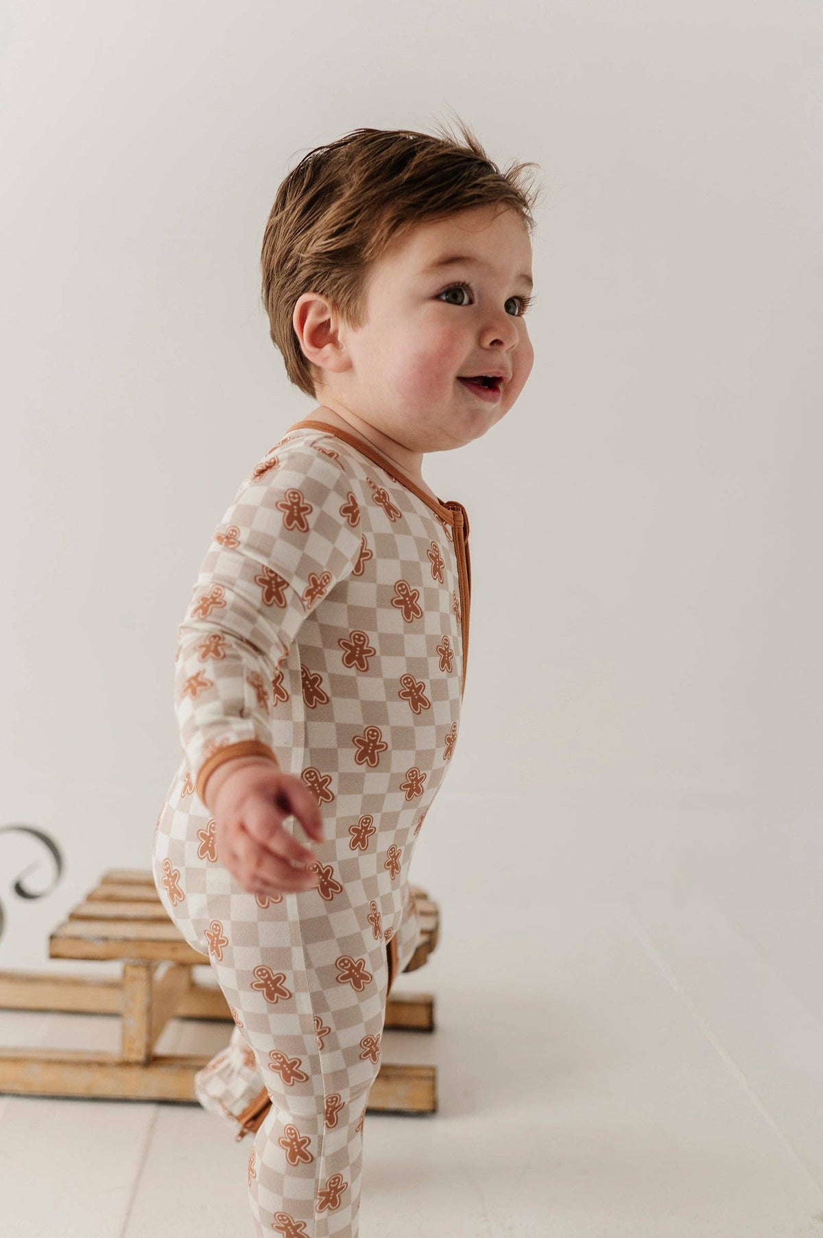 A baby wearing a long-sleeve footed onesie with a beige and white checkerboard pattern and small brown gingerbread motifs, standing in a studio setting.