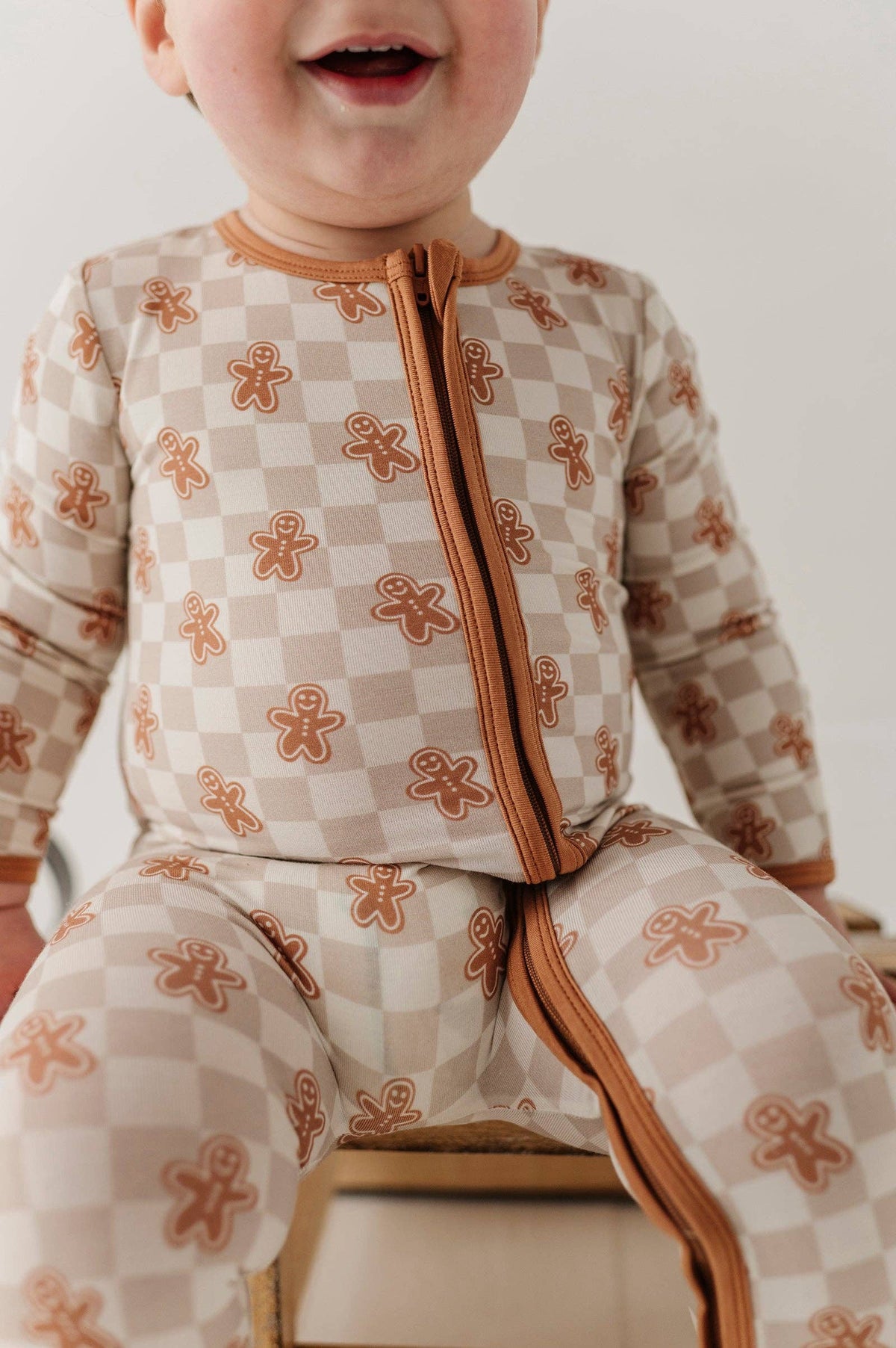 A close-up of a baby wearing a one-piece zip-up sleeper with a beige and white checkerboard pattern and brown gingerbread man motifs, sitting on a wooden stool.