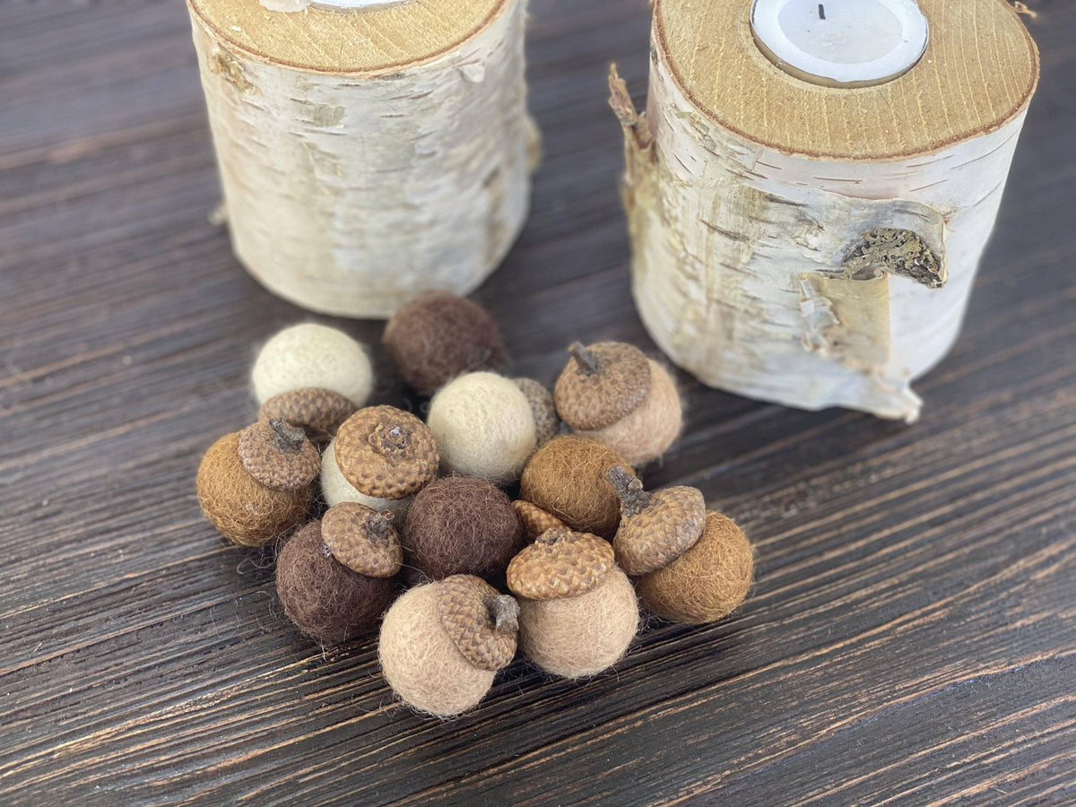 A set of natural acorn-shaped felt candle holders in shades of brown, beige, and cream, displayed on a dark wood surface with two birch log candle holders in the background.