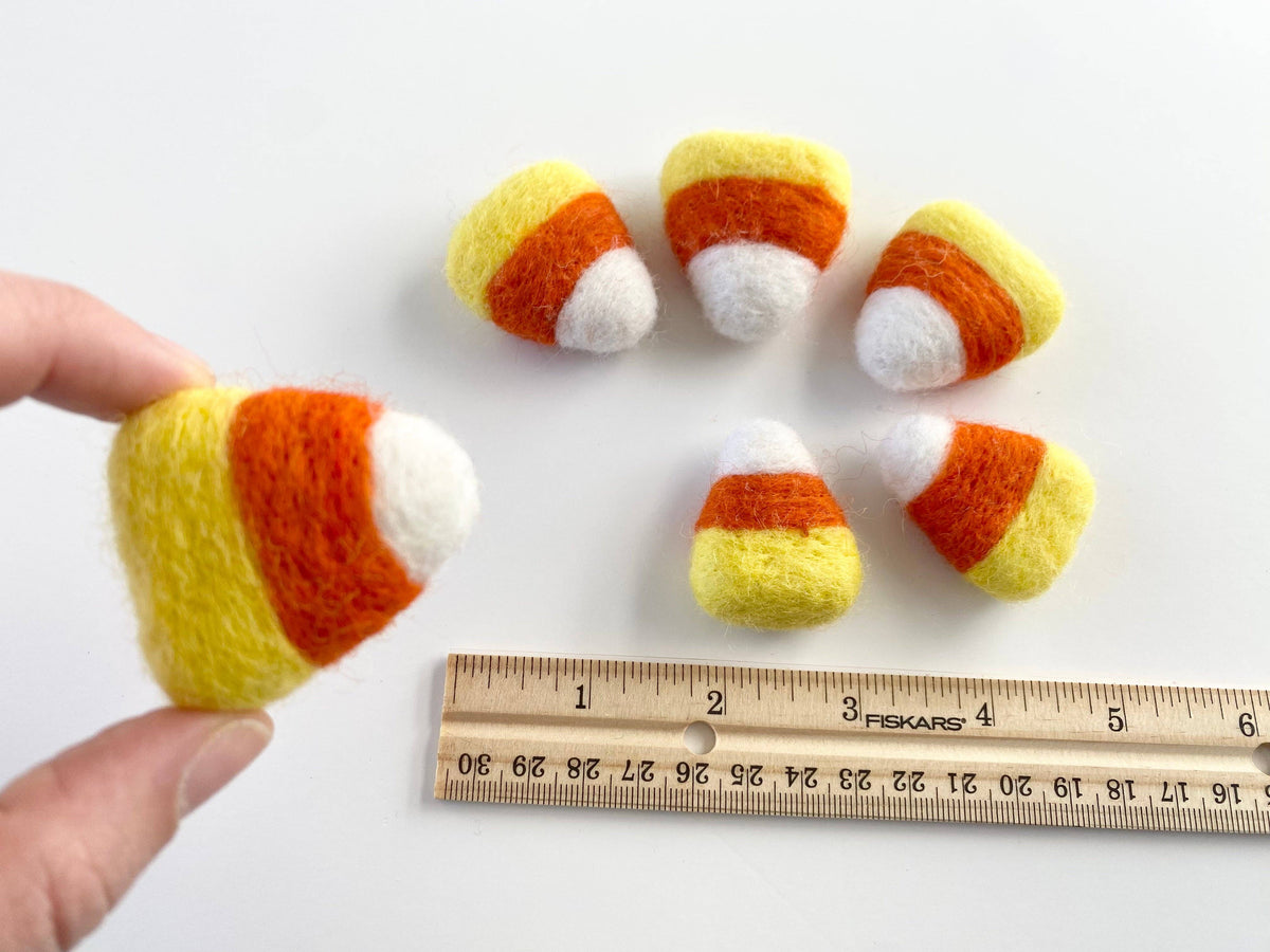 Handmade felted candy corn toys in yellow, orange, and white, with one held by a hand and others arranged on a white surface next to a measuring ruler.