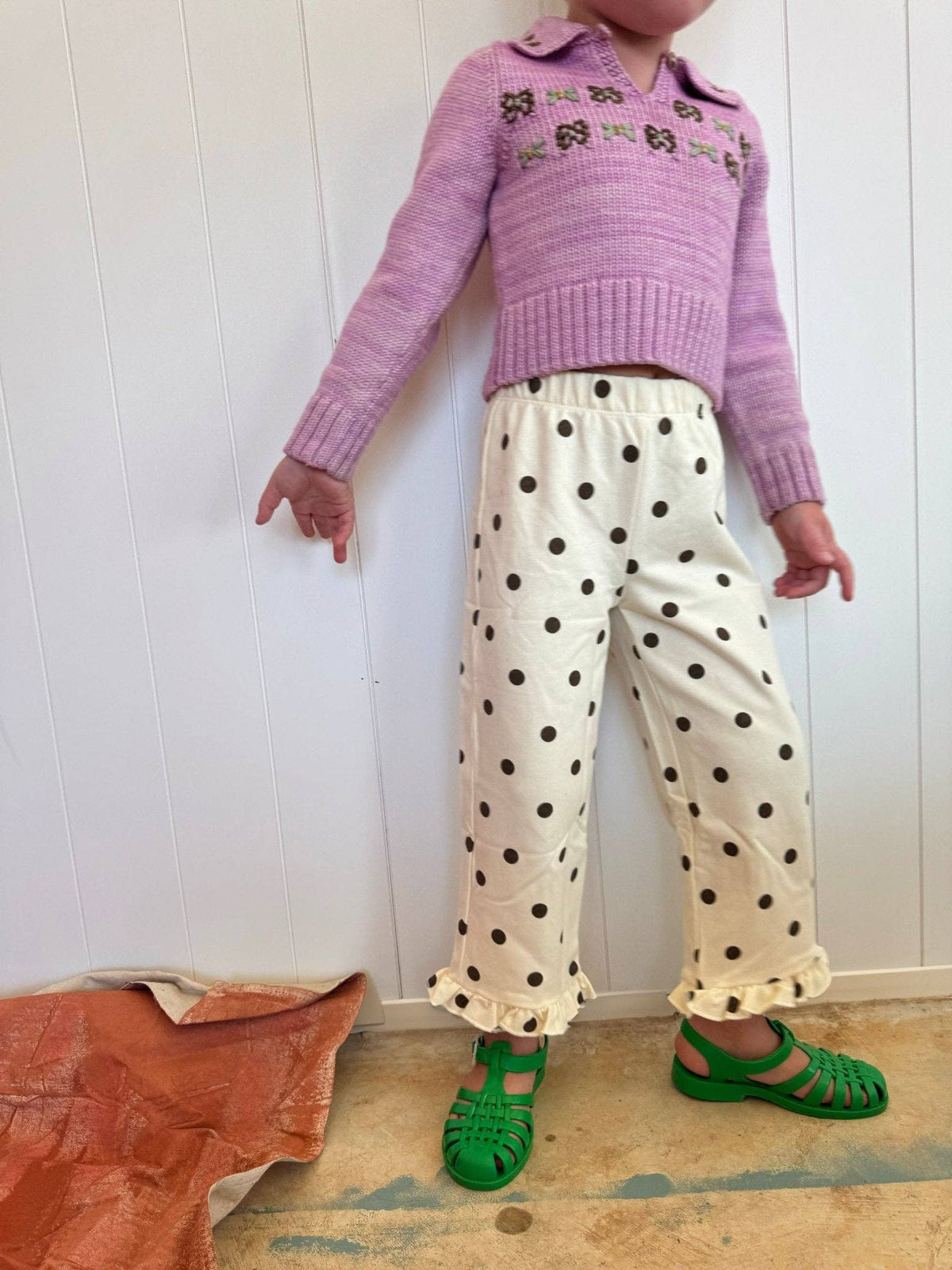A child wearing a lavender knit polo sweater with green floral embroidery, cream wide-leg trousers with black polka dots and ruffled hems, and bright green sandals, standing against a white paneled wall.