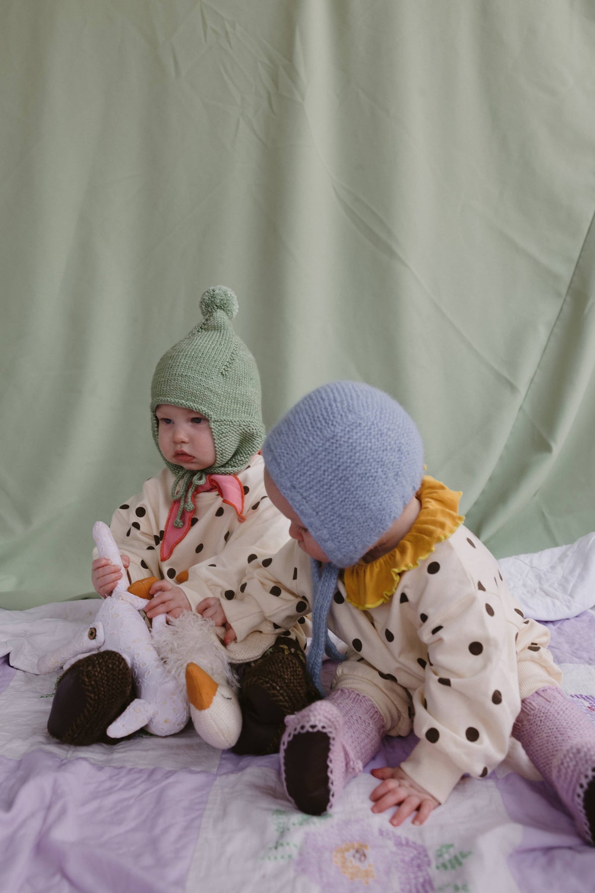 Two toddlers sitting on a lavender quilted bed against a sage green fabric backdrop. The child on the left wears a light green knit bonnet with ear flaps, a cream polka-dot jacket, and dark brown boots while holding a white plush duck.