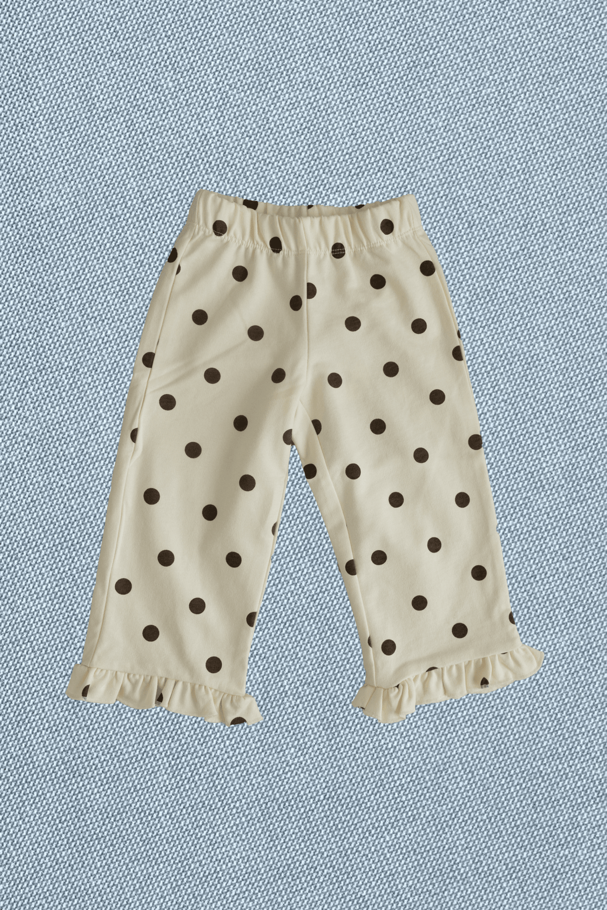Cream-colored toddler pants with a black polka dot pattern and ruffled hems, laid flat on a textured blue background.