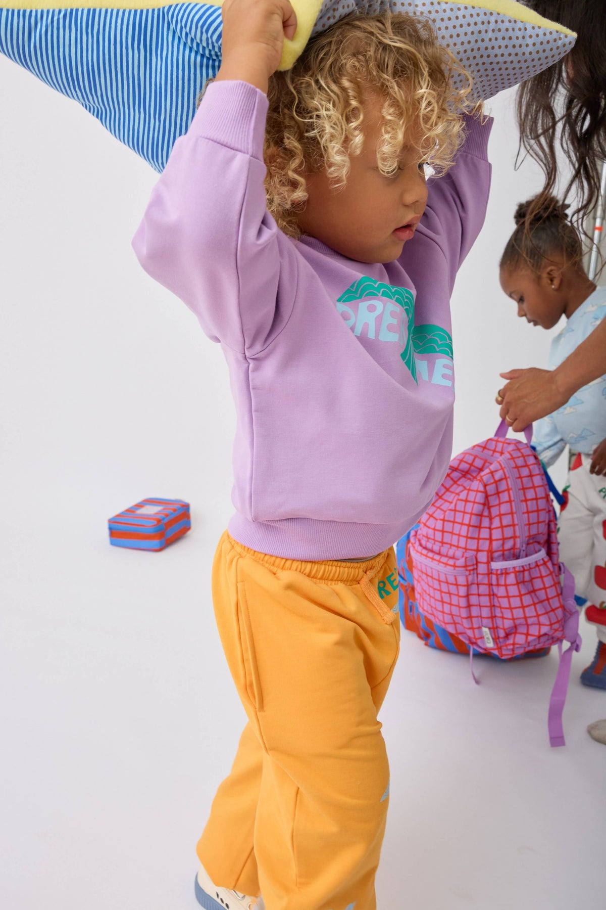 A toddler wearing a lavender sweatshirt and bright yellow pants stands in a studio setting, holding a striped blue and yellow pillow overhead. Another child and a pink backpack are visible in the background.