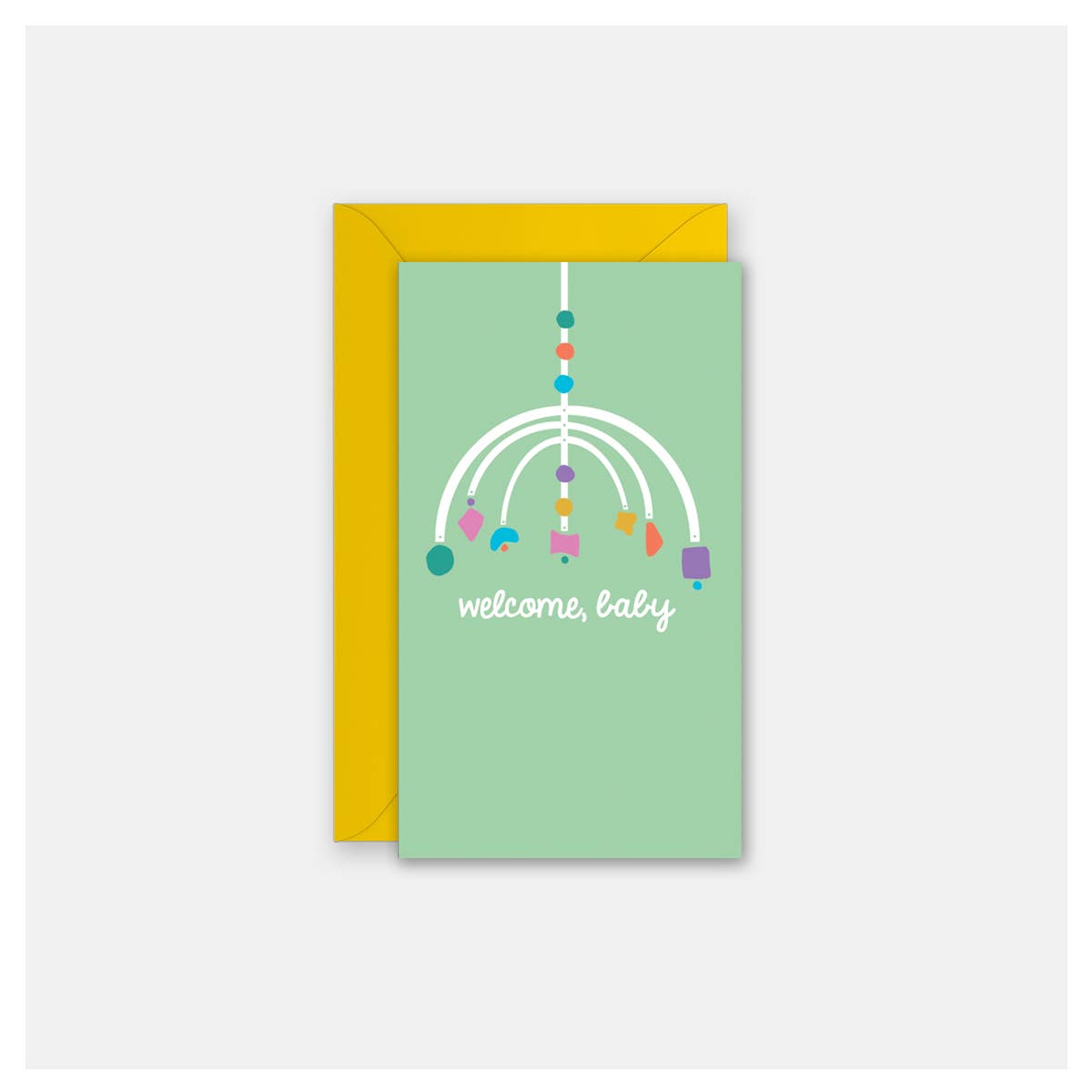 A baby welcome card with a light green front featuring a white mobile graphic and the text 'welcome, baby' in white script, shown with a yellow envelope behind it on a light gray background.