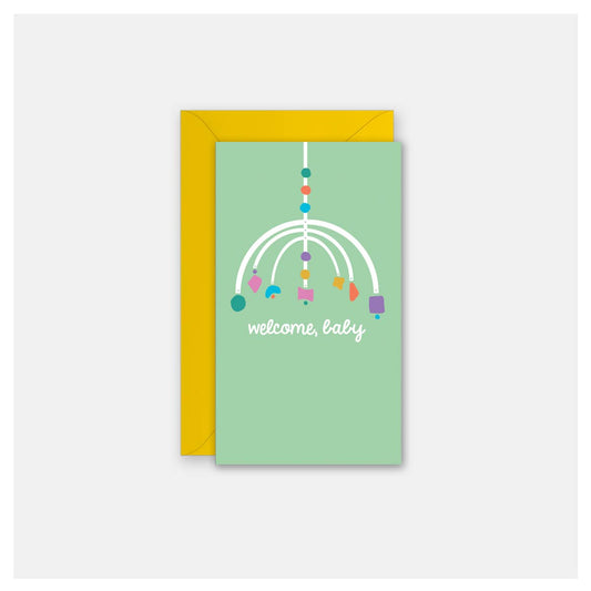 A baby welcome card with a light green front featuring a white mobile graphic and the text 'welcome, baby' in white script, shown with a yellow envelope behind it on a light gray background.