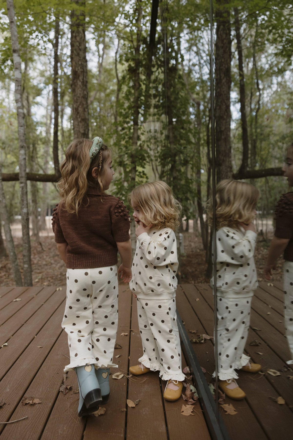 Three toddlers standing on a wooden boardwalk in a wooded outdoor setting, wearing matching outfits. The outfits consist of a dark brown knit top and white pants with black polka dots, paired with light blue rain boots and tan shoes.