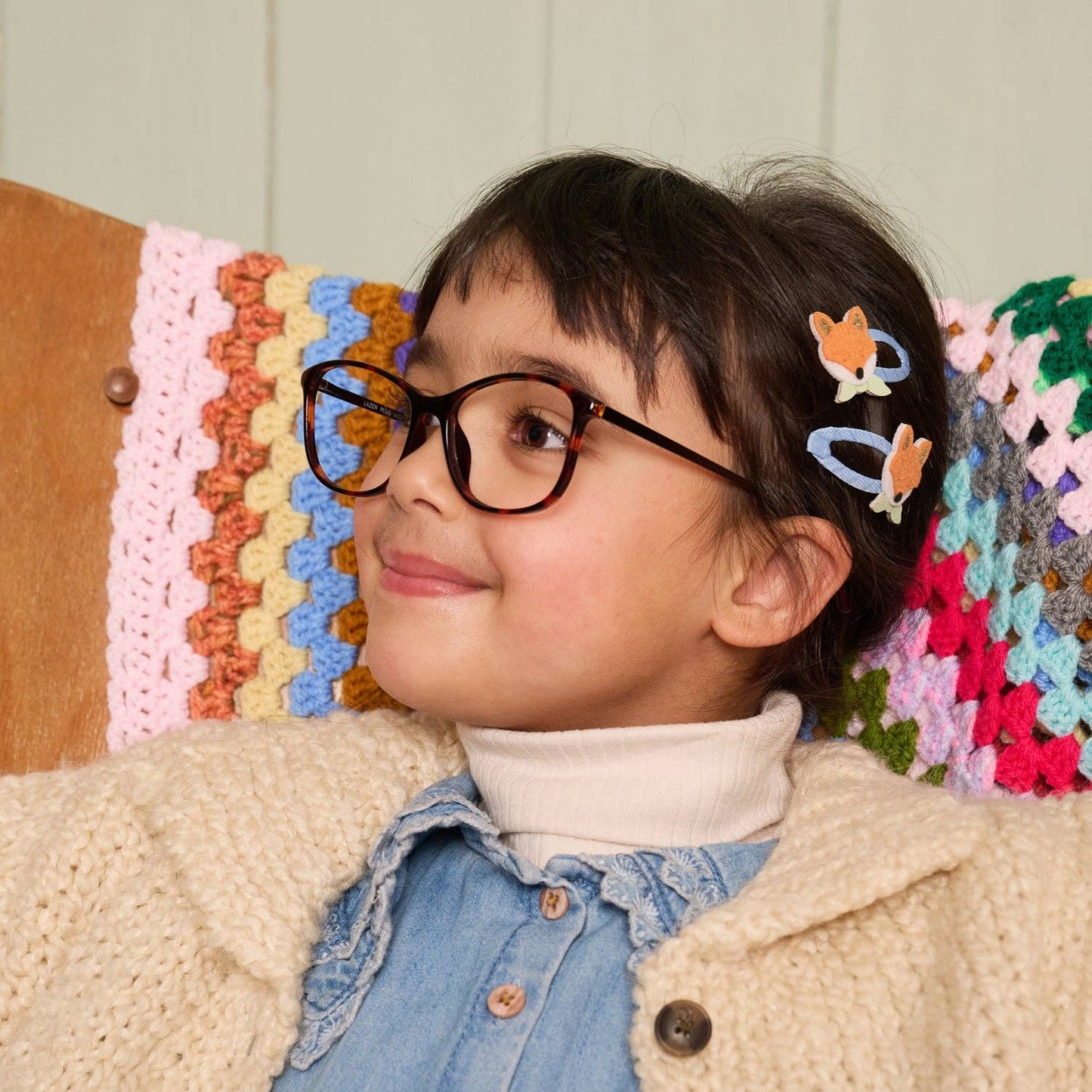 A young girl with dark hair wearing black-rimmed eyeglasses and two butterfly-shaped hair clips, smiling while looking to the side. She is wearing a cream cardigan over a light blue denim shirt and white turtleneck, sitting in front of a colorful crocheted blanket.