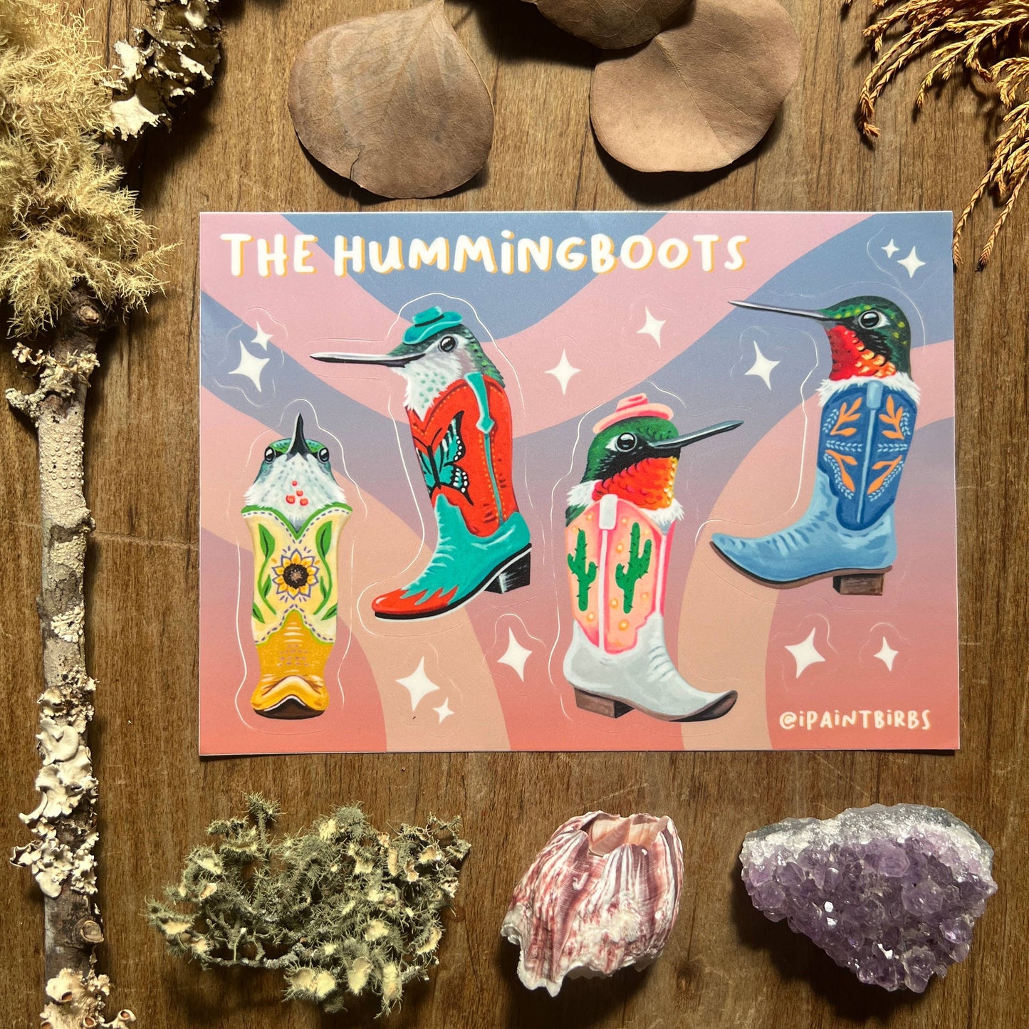 Card with colorful illustrations of boots and hummingbirds on a wooden surface with decorative elements.