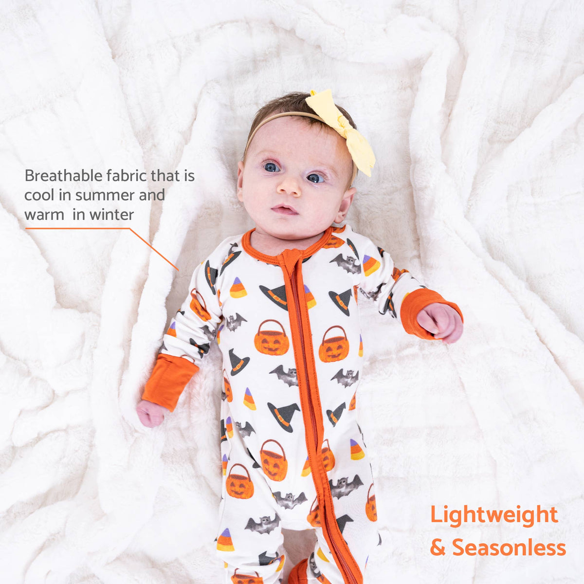 A baby lying on a white textured blanket wearing a white zip-front footed sleeper with orange trim and a Halloween pumpkin, witch hat, and candy corn print. Text on the image highlights breathable fabric and lightweight seasonless design.