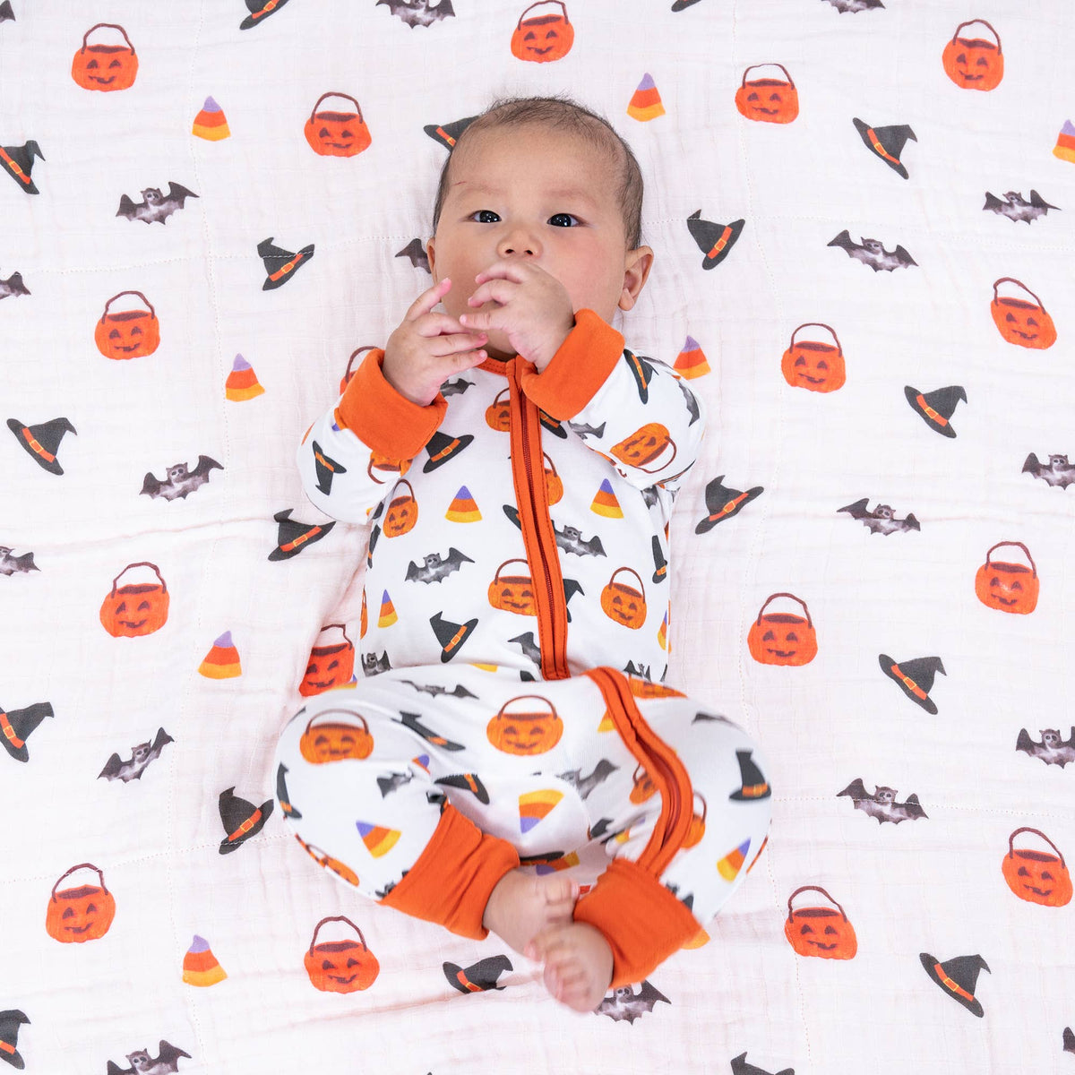 A baby lying on a white muslin blanket with an orange, black, and yellow Halloween print, wearing a matching zip-up footed sleeper with orange cuffs and collar.