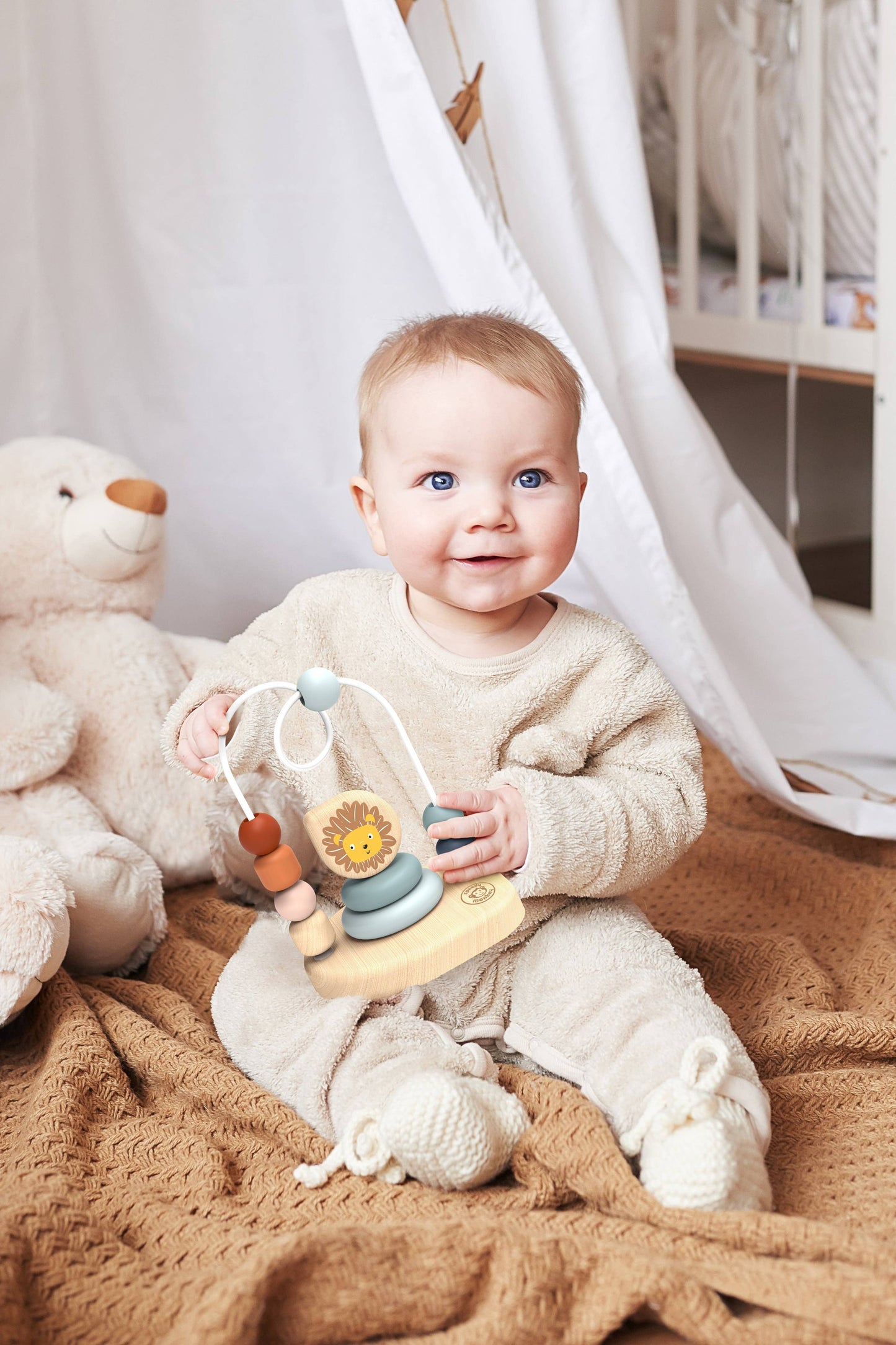 Baby sitting on a blanket holding a wooden toy with a teddy bear and white curtains in the background.