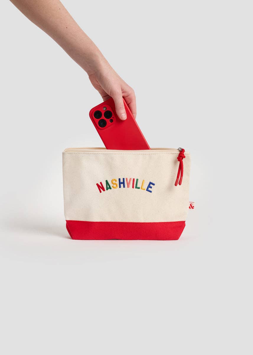 A hand holding a red smartphone above a cream canvas zip pouch with a red bottom panel and colorful 'NASHVILLE' text on the front, set against a light gray studio background.