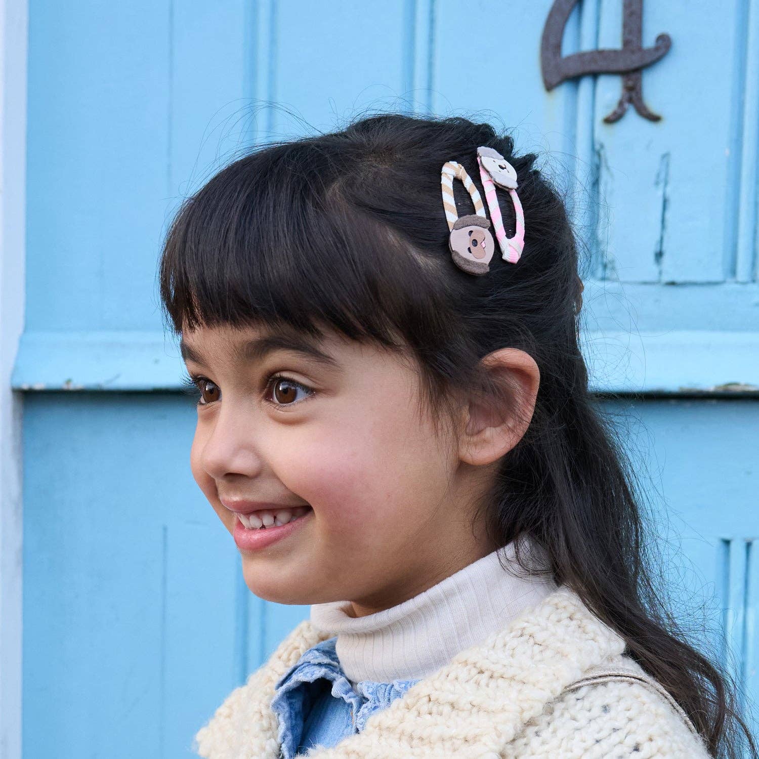A young girl with dark hair wearing a pink and white decorative hair clip, shown in profile against a light blue wooden door.