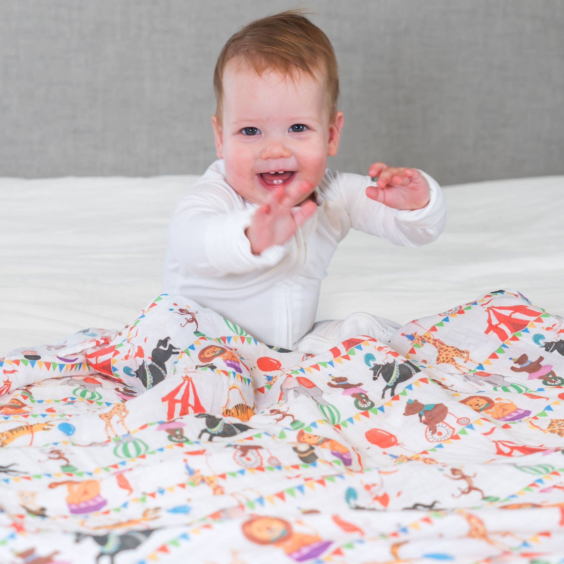 Baby lying on a colorful blanket with animal patterns, smiling and reaching out.
