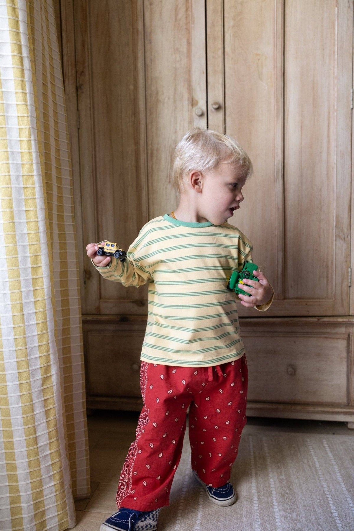 A toddler standing indoors in front of a wooden wardrobe, wearing a pale yellow and green striped long-sleeve top, red pants with small white motifs, and blue sneakers, while holding small toy objects in both hands.