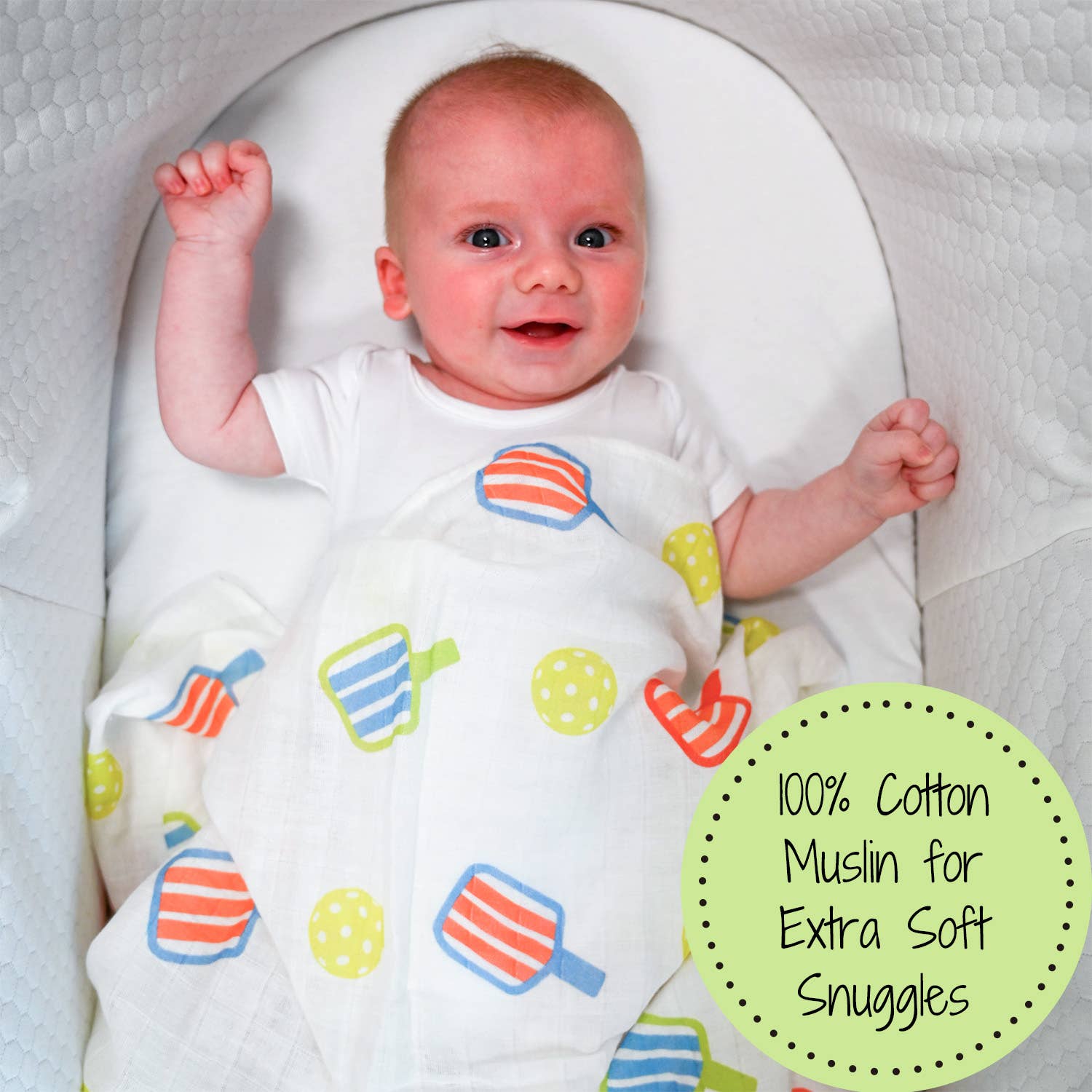 Baby lying on a white cushion wearing a colorful baby wrap with a label indicating 100% cotton muslin.