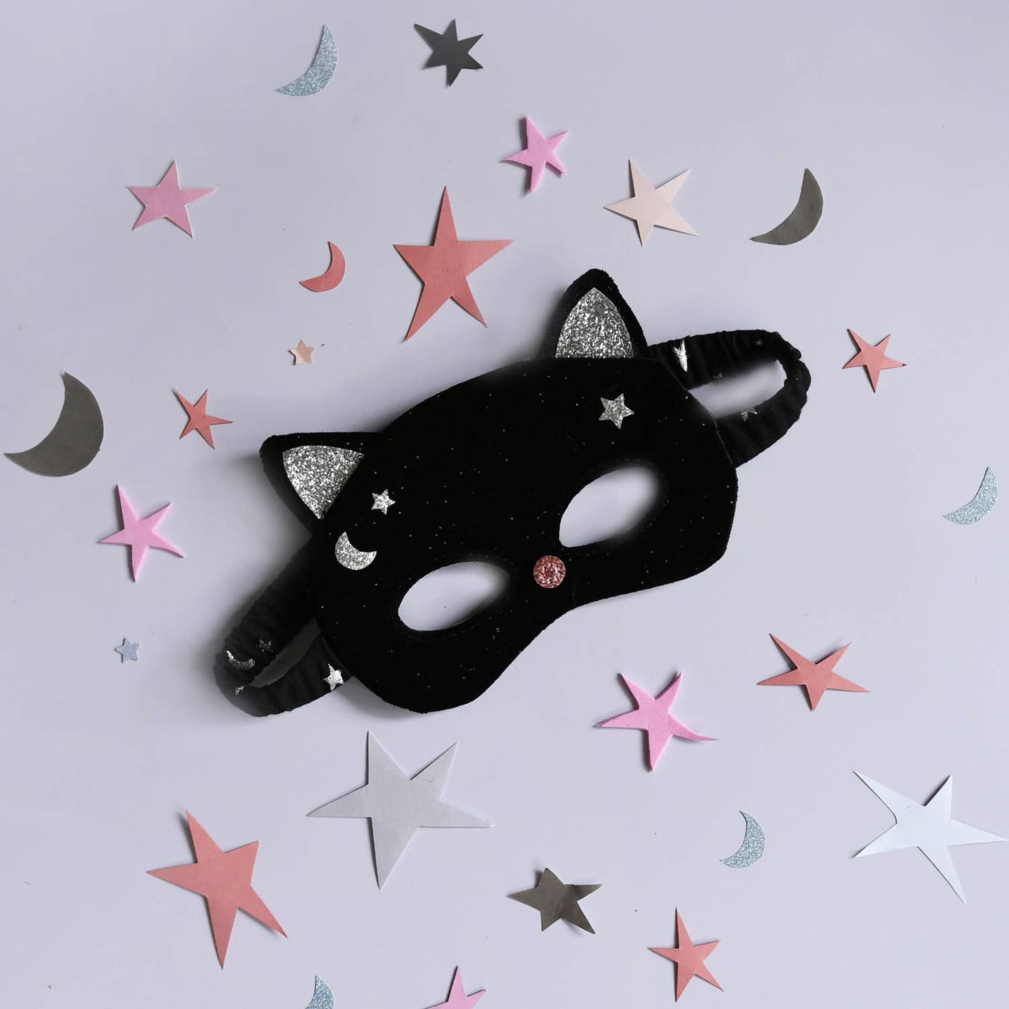 A black cat-themed party mask with glittery silver ears and stars, decorated with a small pink rhinestone, shown on a light gray background with scattered pink, white, and silver star and moon confetti.