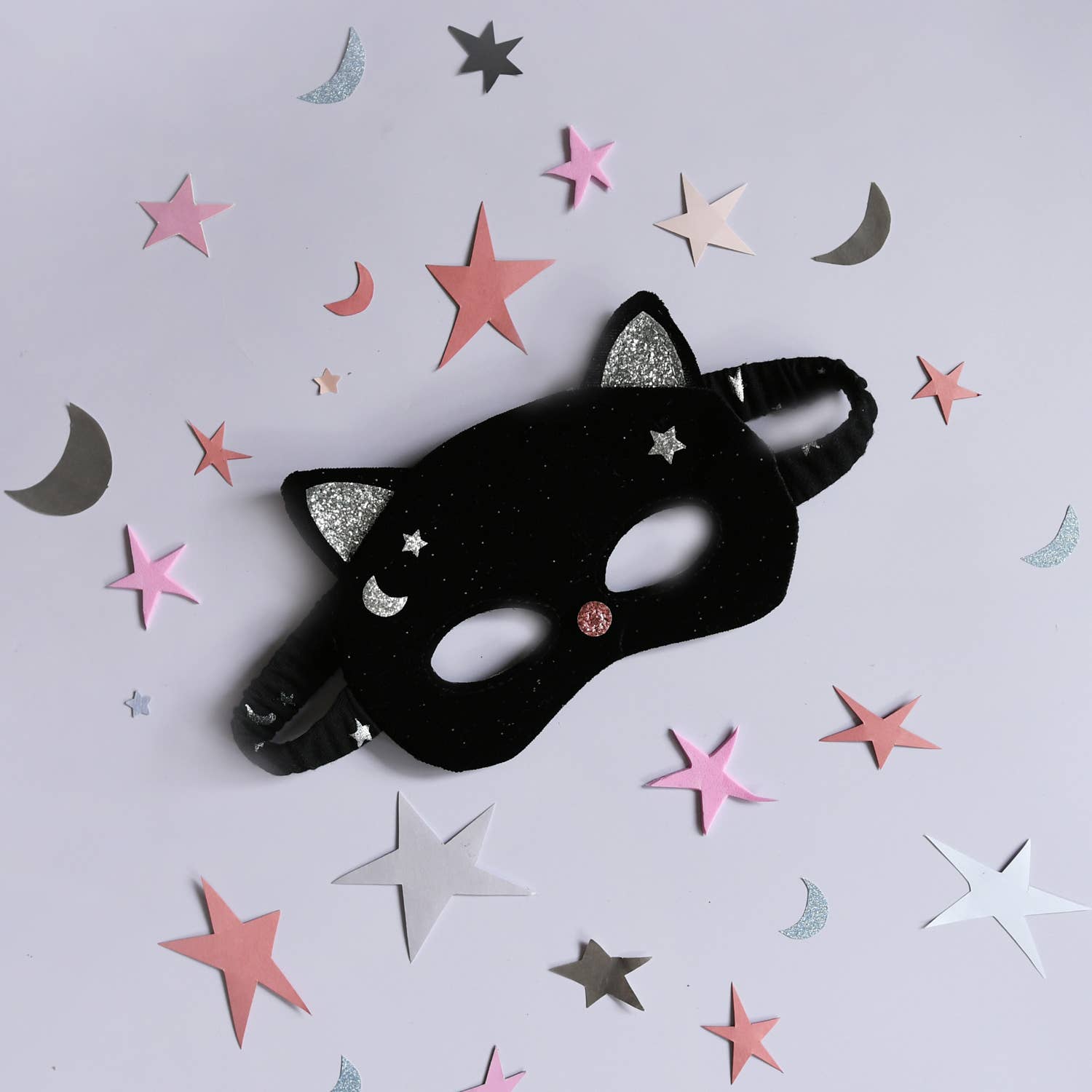 A black cat-themed party mask with glittery silver ears and stars, decorated with a small pink rhinestone, shown on a light gray background with scattered pink, white, and silver star and moon confetti.